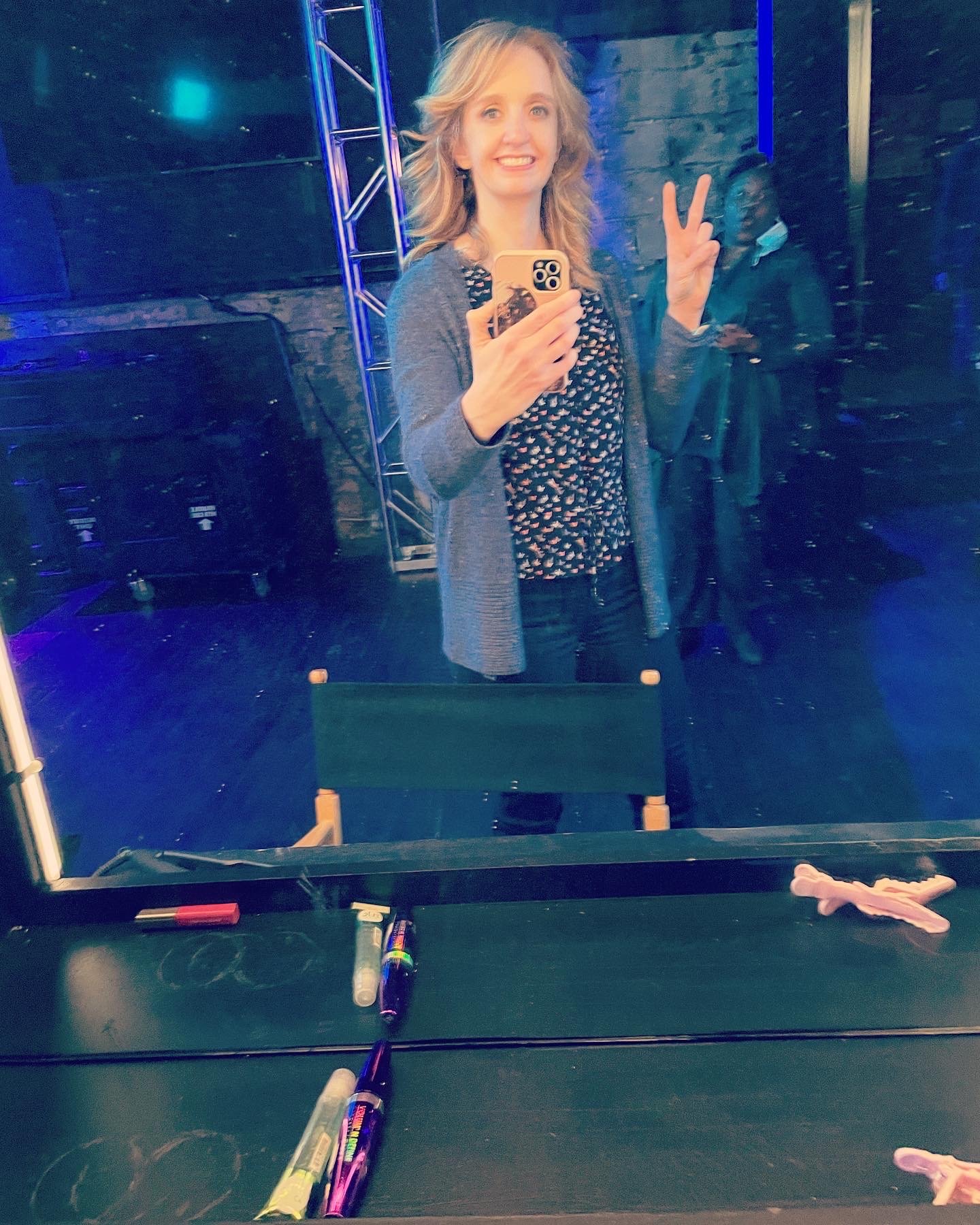 A woman with shoulder-length blonde hair taking a selfie in a mirror, smiling and making a peace sign with her right hand. She wears a patterned blouse and a gray cardigan, standing in a dimly lit room with blue and purple lighting. There are various