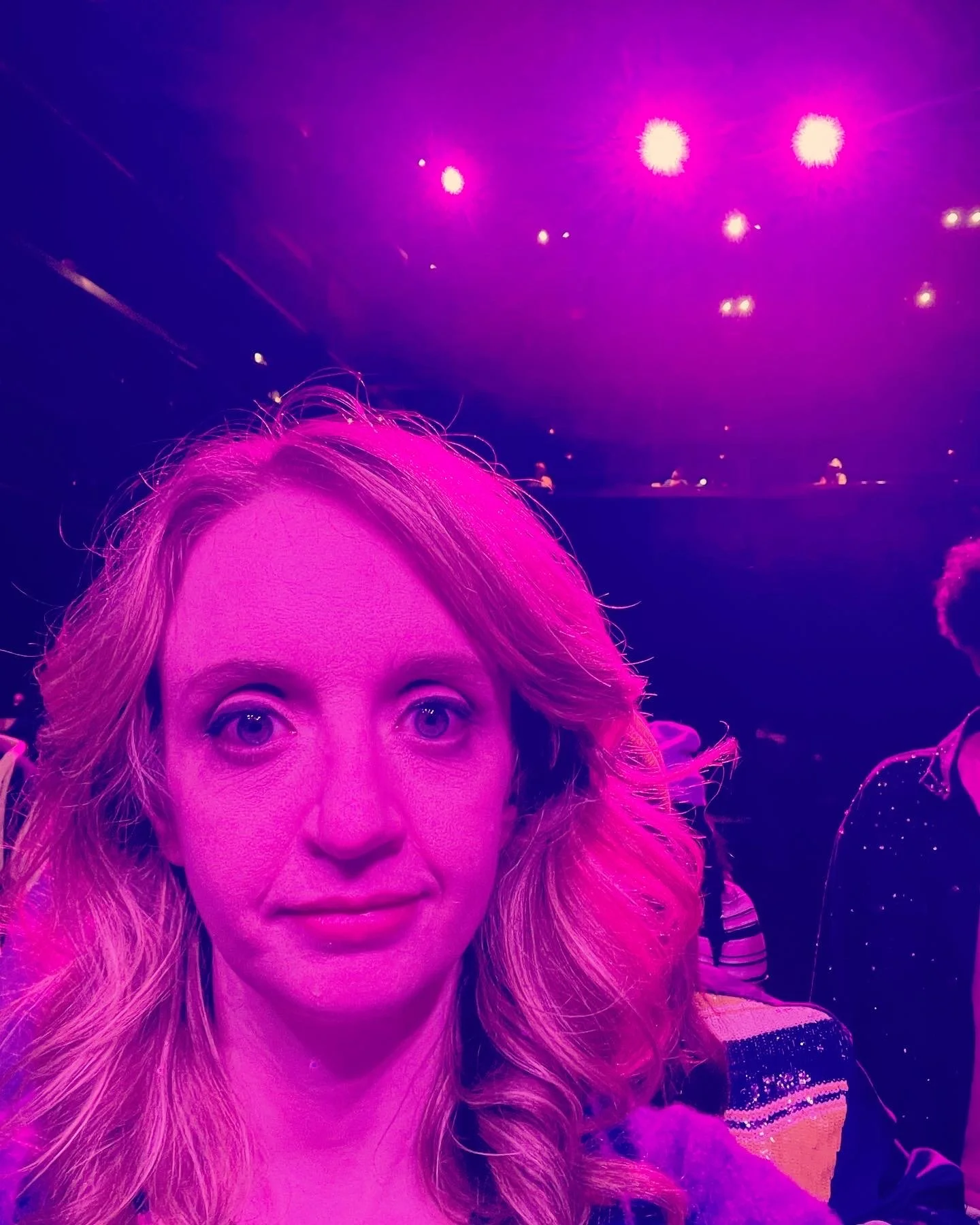 Close-up of a woman with blond hair under purple and pink lighting at an indoor event or concert.