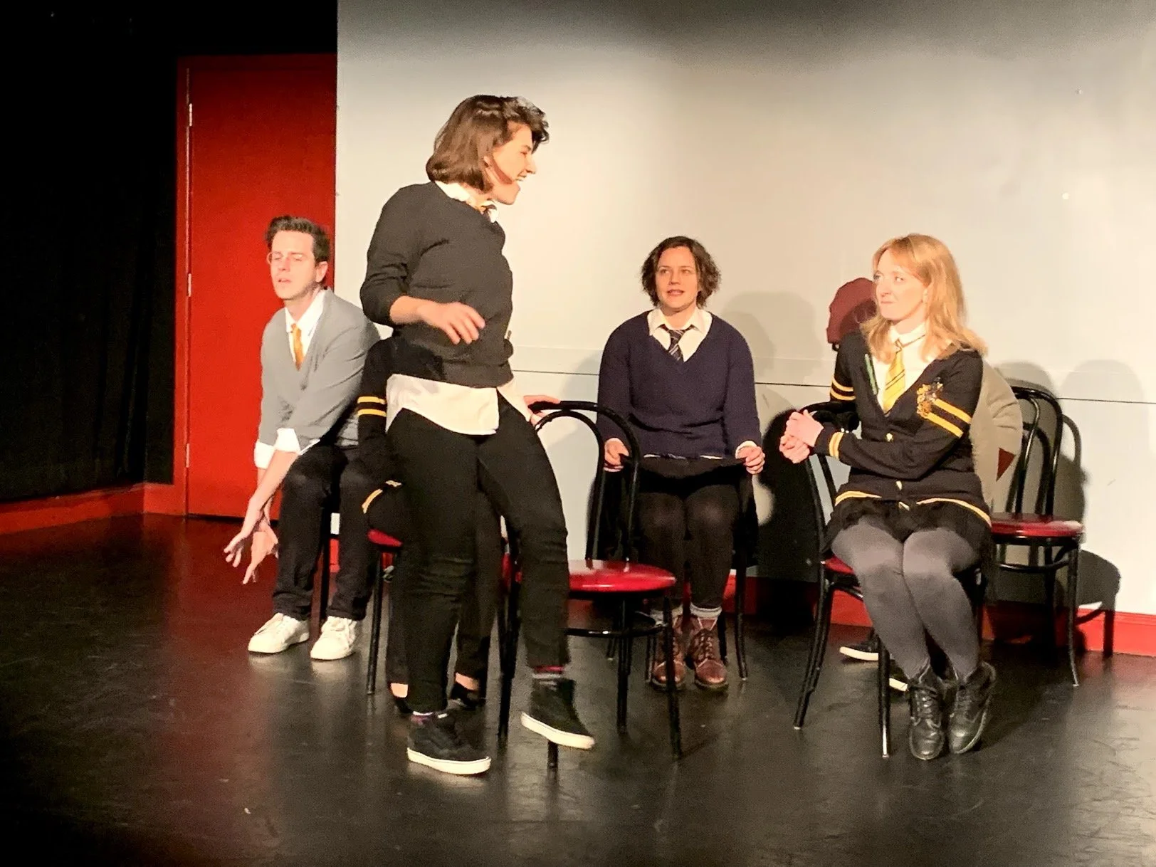 Four actors on stage, three are sitting on chairs and one is standing, during a theatrical performance, with a white and red background.