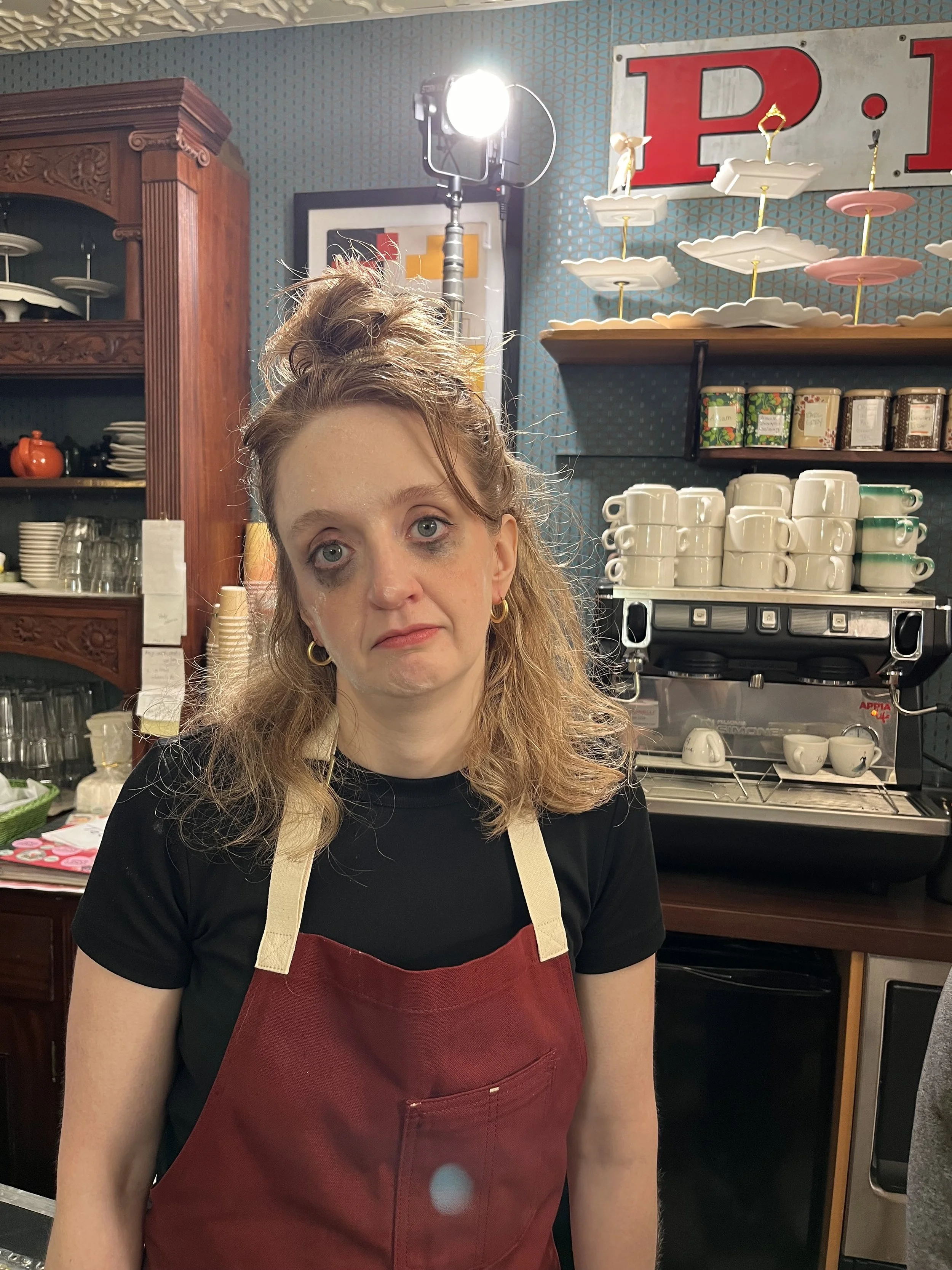 A woman with curly hair, wearing a red apron, standing in a kitchen with shelves of cups, dishes, and tins behind her. There is a coffee machine on the counter.