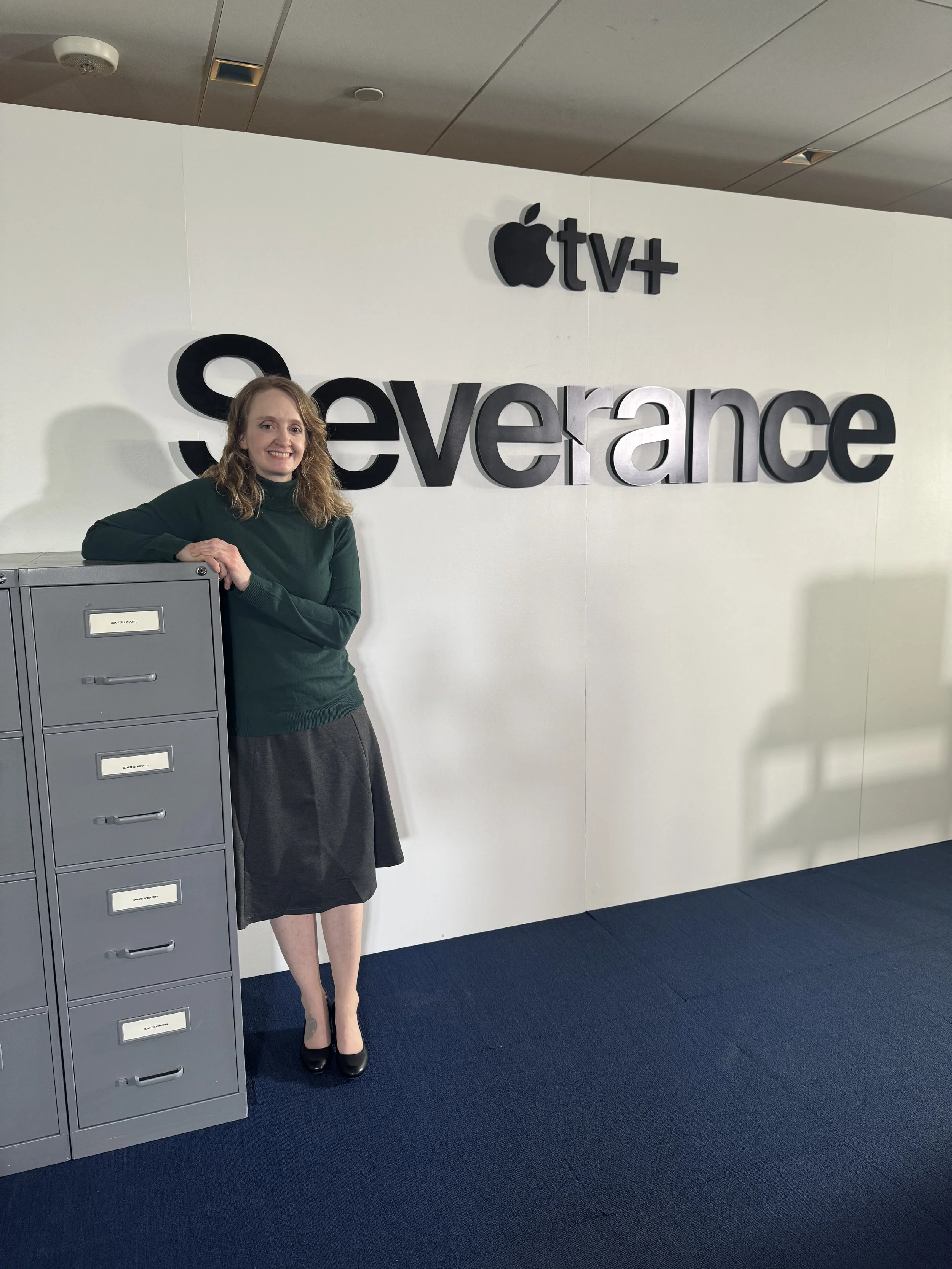 A woman with blonde hair, wearing a dark green sweater and black heels, standing near a gray metal locker in front of a white wall with black three-dimensional sign letters that say "Severance" and the Apple TV+ logo above.