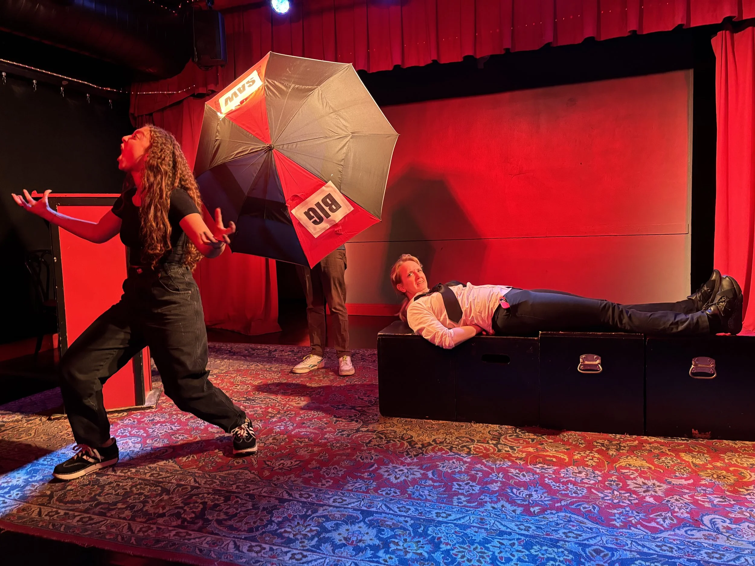 Stage performance with four actors on stage; two are standing, one sitting, and one lying on a bench. The stage has a red curtain background, a dark-colored floor, and a vibrant patterned carpet.