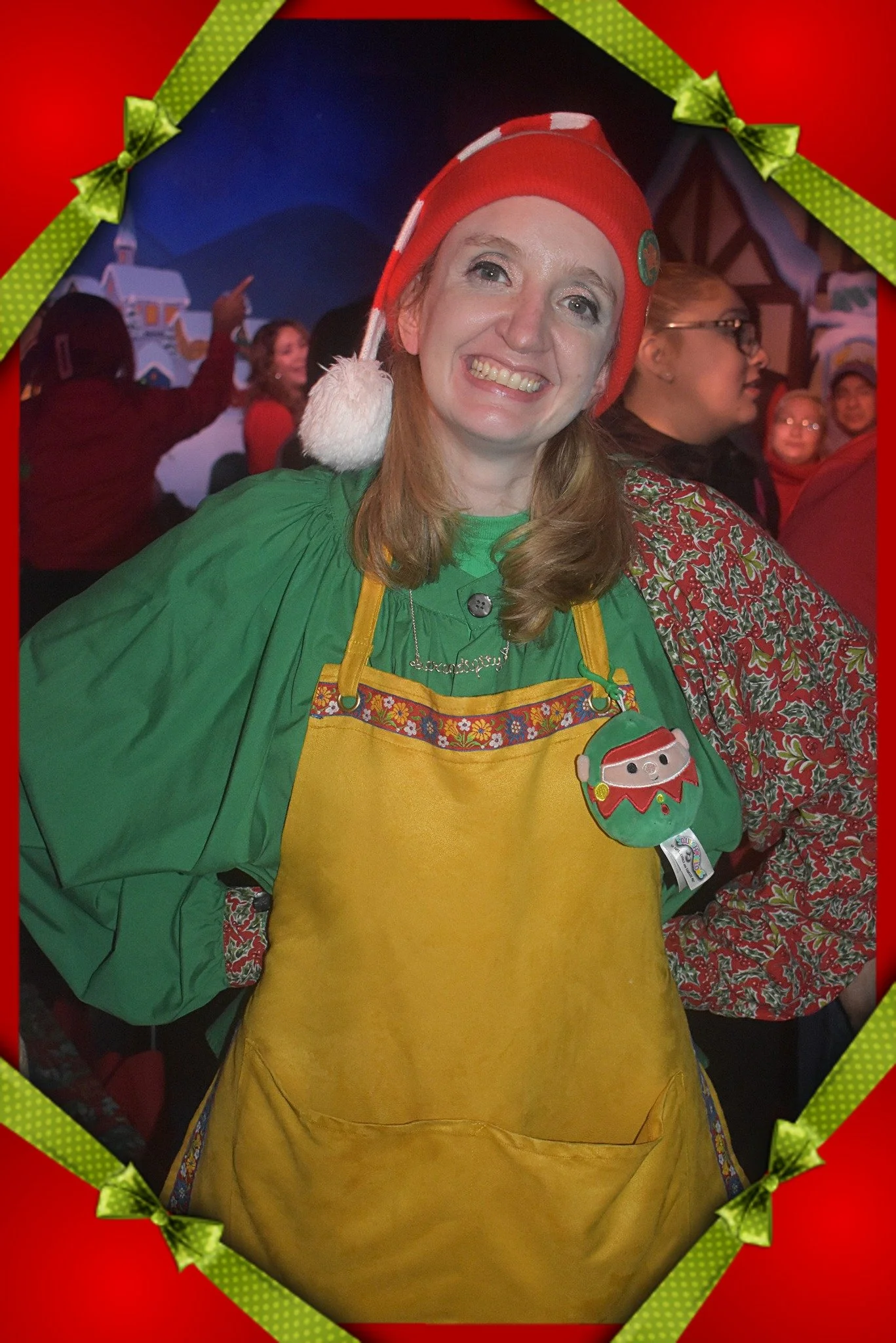 Woman dressed as an elf with a red hat and green shirt, smiling at a holiday gathering, holding a stuffed Santa doll, with Christmas decorations and other people in the background.