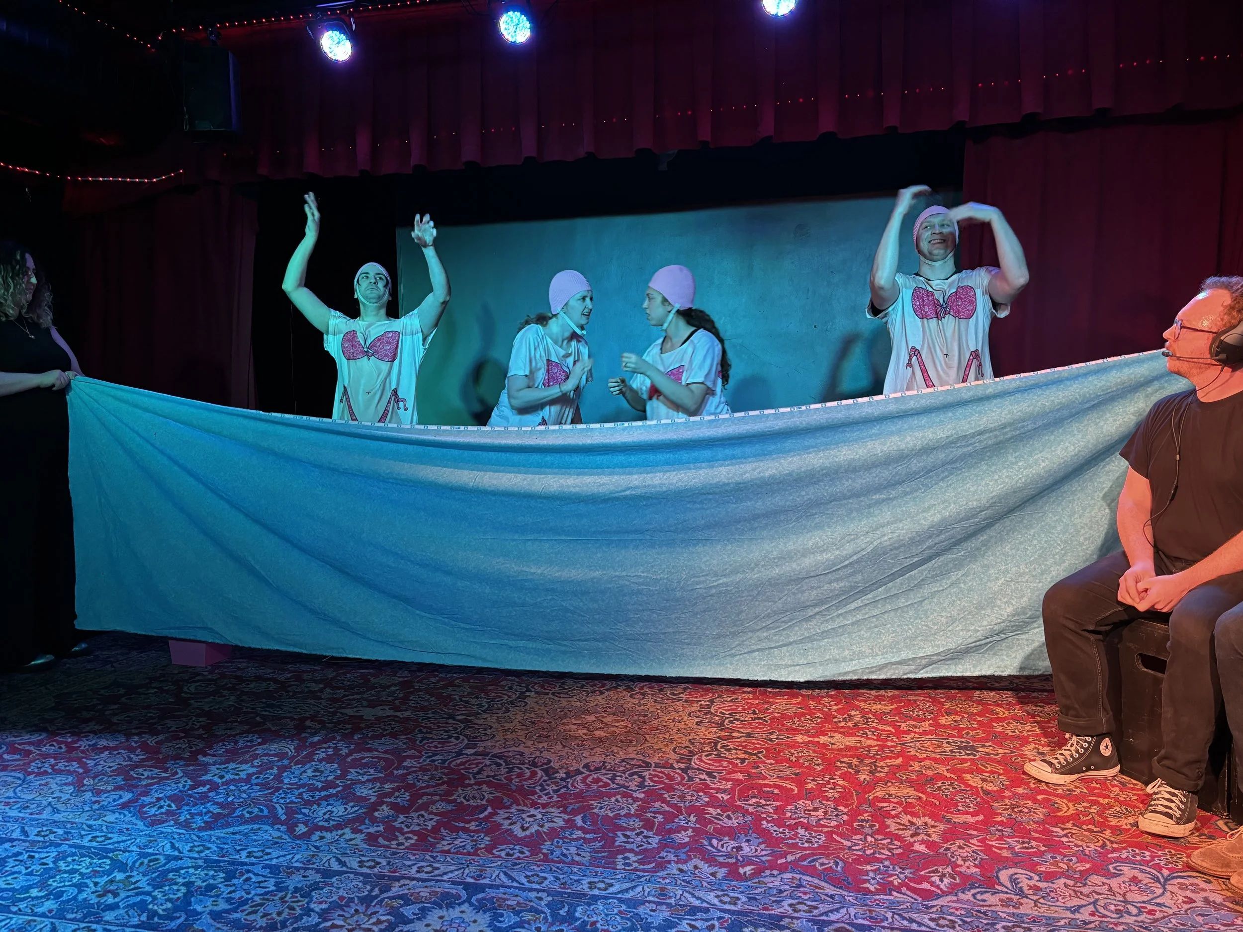 People on stage dressed in matching T-shirts and hats, performing a dance or act under colorful stage lights, with a person sitting in the foreground.