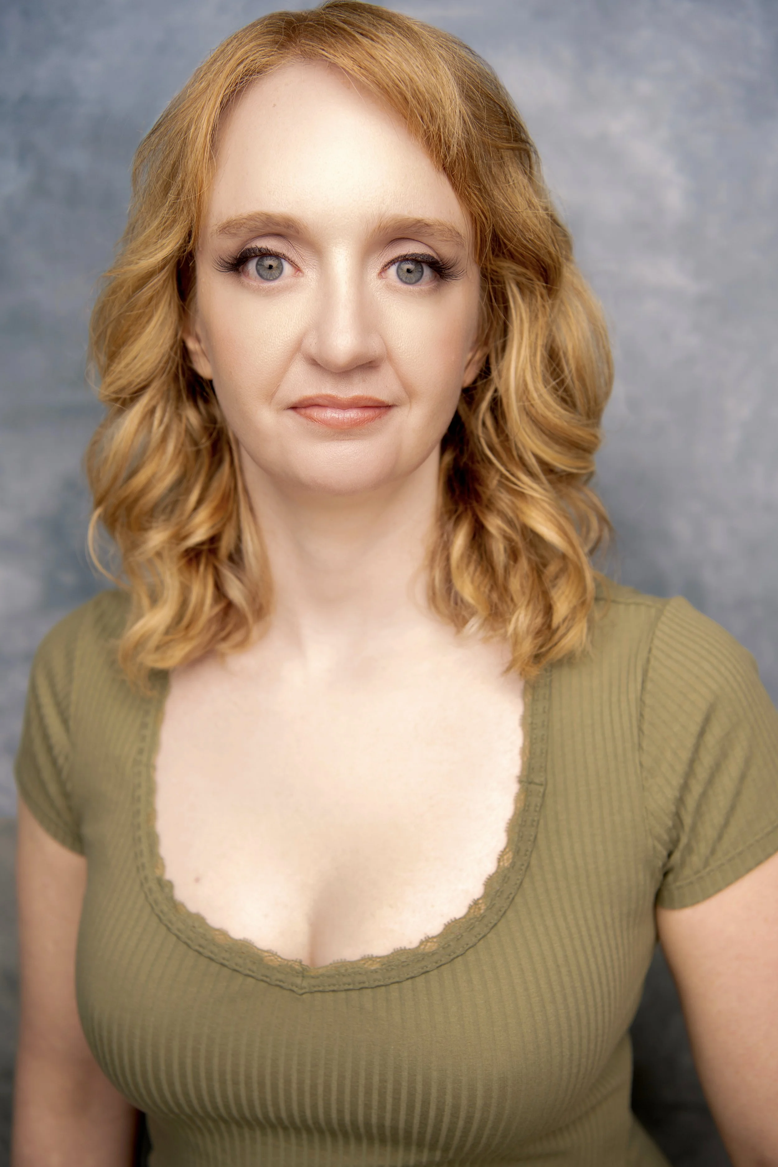 Close-up of a woman with curly red hair and blue eyes, wearing a green top with a lace neckline, against a bluish-gray background.
