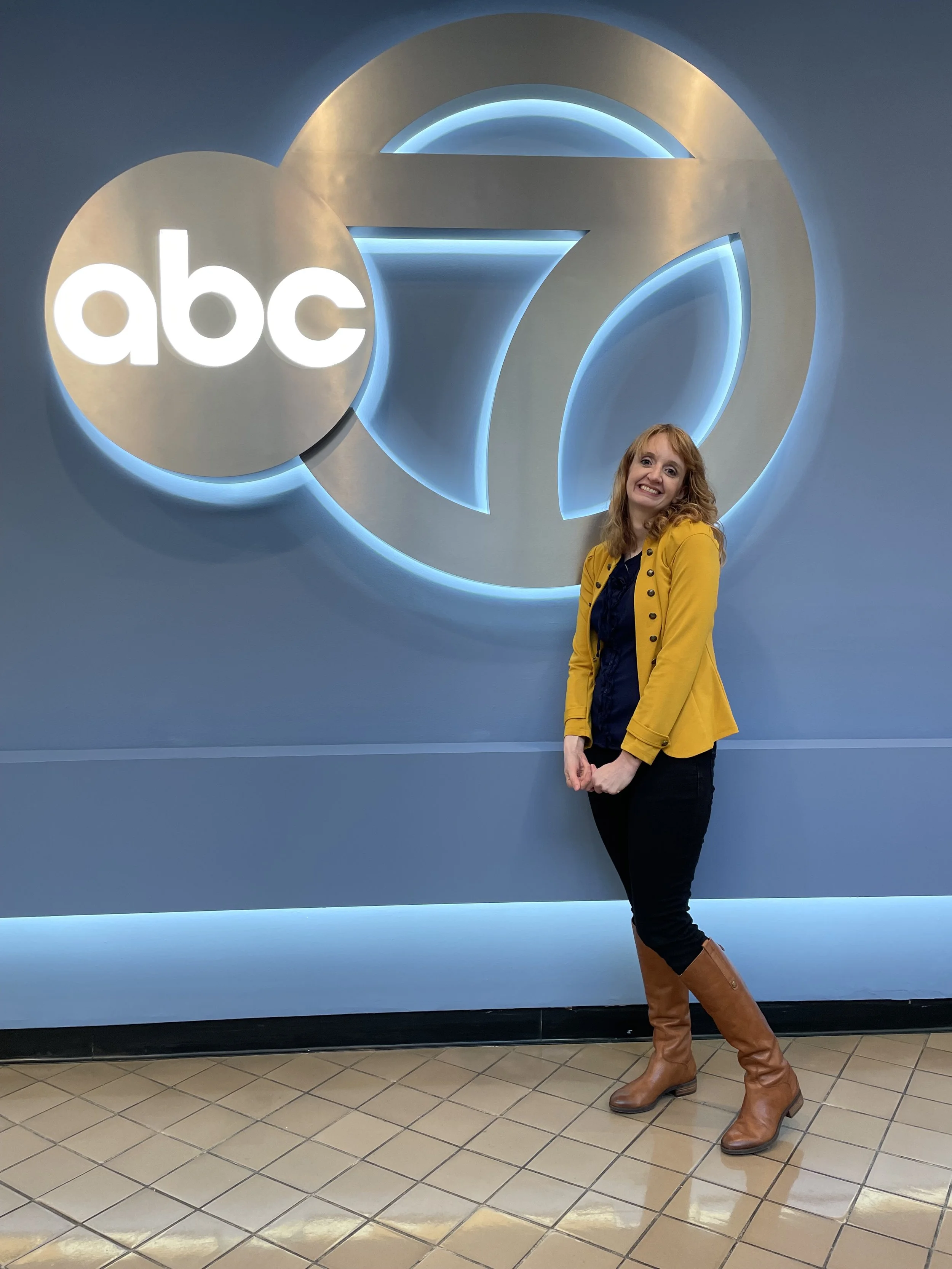 A woman in a yellow jacket, black pants, and brown knee-high boots smiling and standing in front of an ABC logo sign with a blue background.