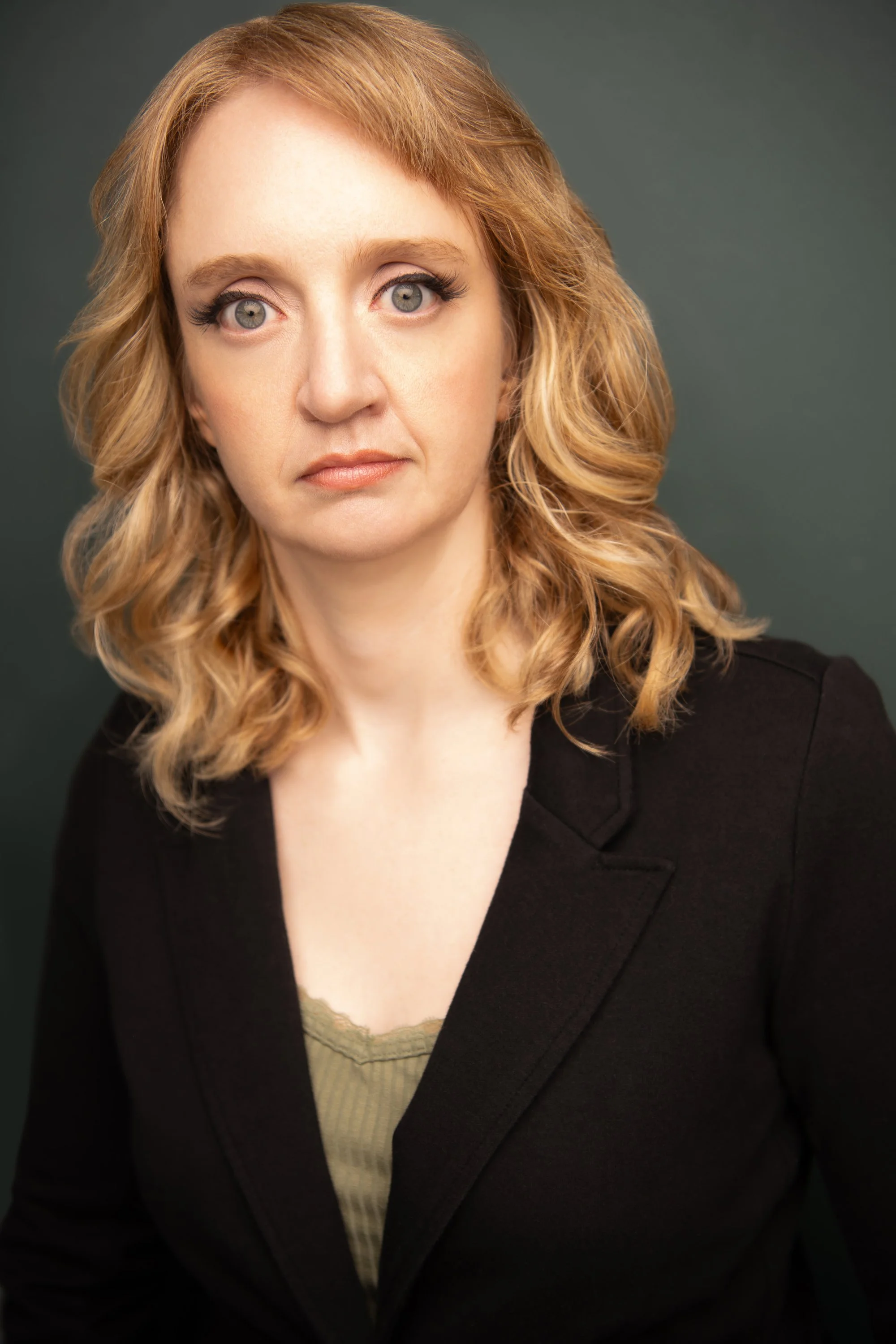 A woman with curly blonde hair and grey eyes wearing a black blazer over a green top.