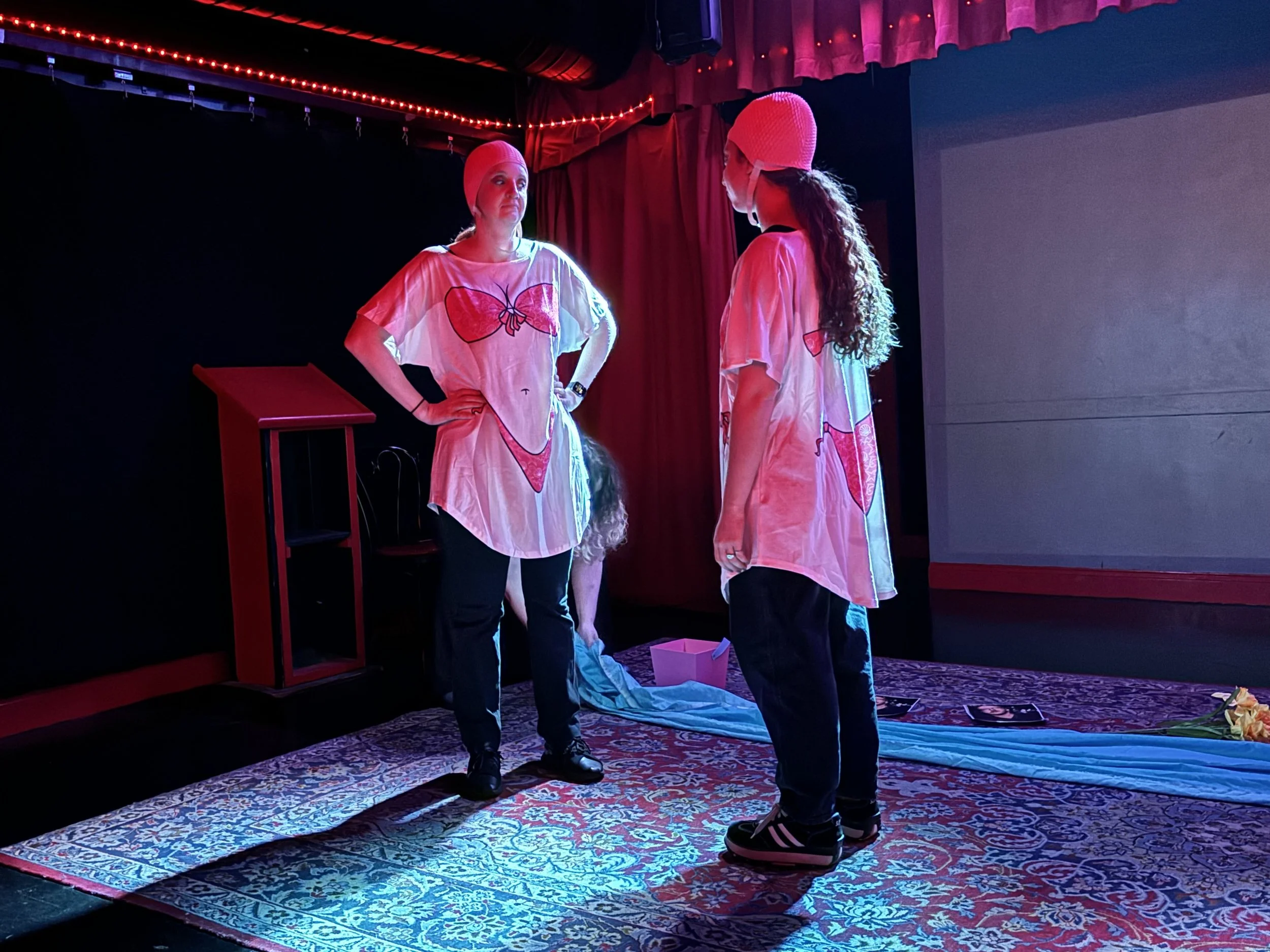 Two women on stage wearing pink hospital gowns with heart and butterfly prints, and pink knit caps, looking at each other, with red curtains and string lights in the background.