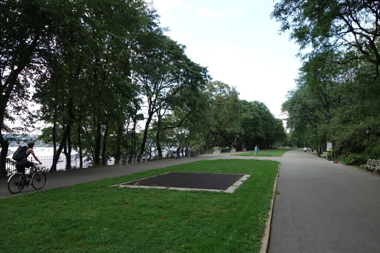 Paving and blocks of mowed grass cover the Serpentine Promenade. Large metal grates allow air to escape from the train tunnel beneath. The 91st Street Community Garden can be seen in the far distance.