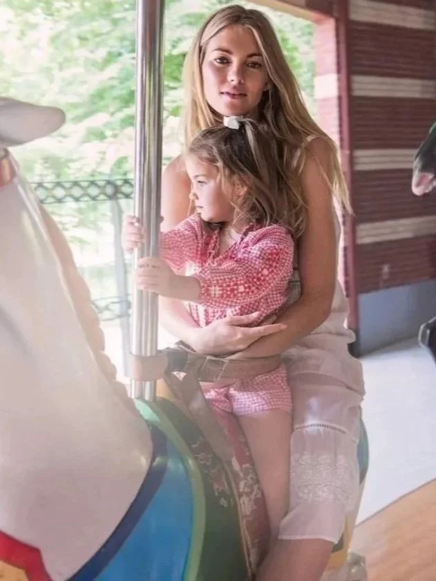 A young woman sitting on a carousel horse while holding a little girl, both smiling inside an amusement park.