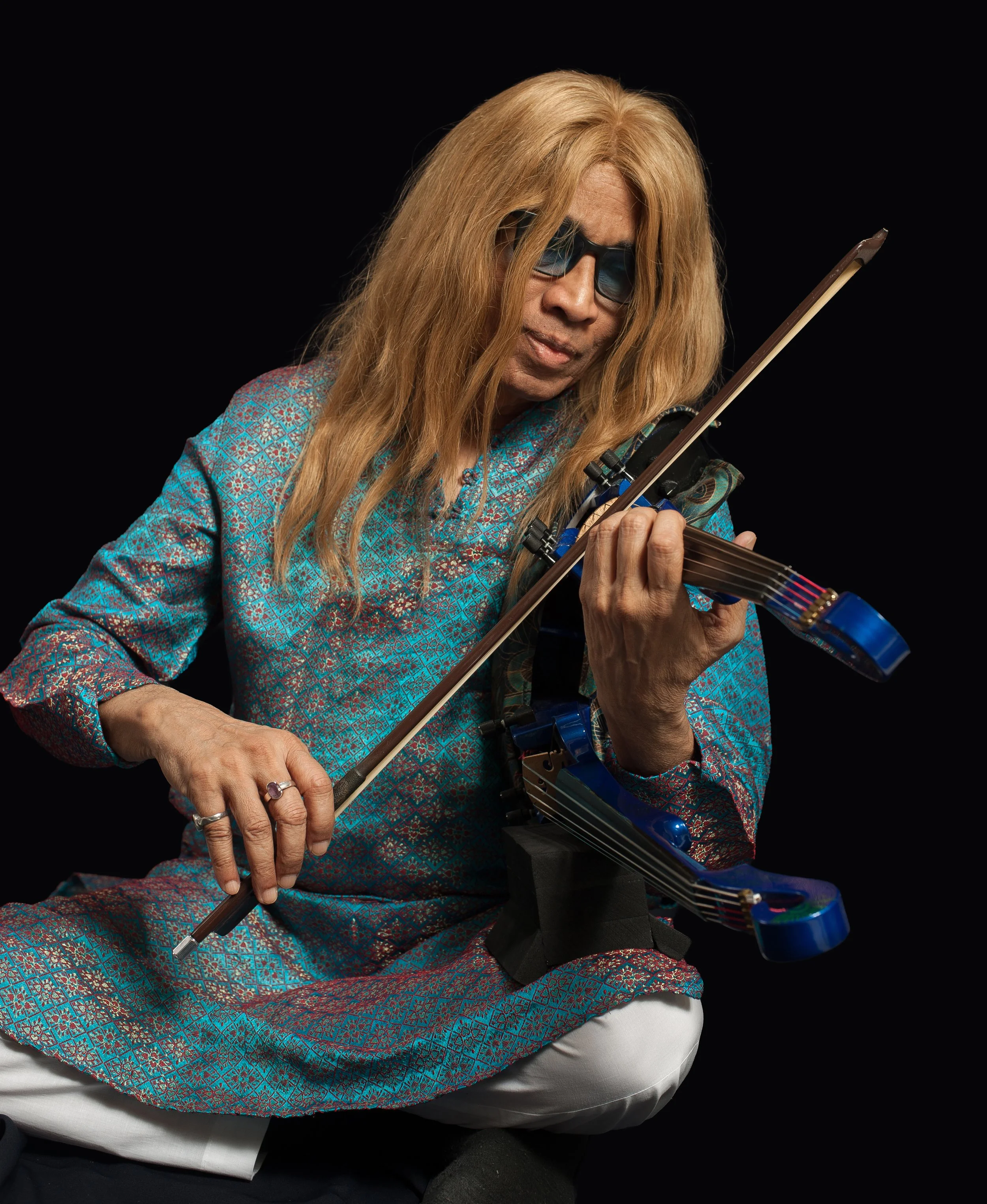 L. Shankar wears sunglasses and a tunic and is seated playing the double violin