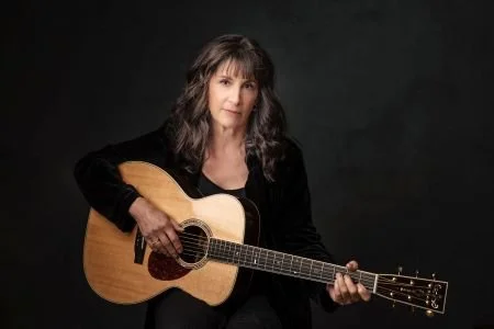 Karla Bonoff is seated, playing guitar and looking straight ahead thoughtfully