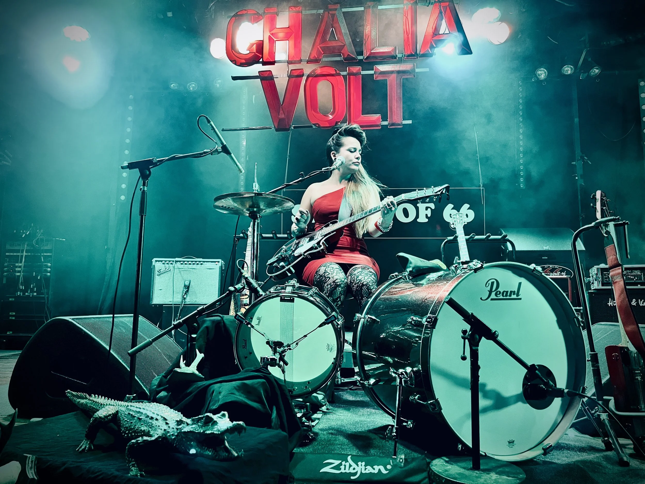Ghalia Volt sits onstage behind a drum kit, playing guitar. A large sign says GHALIA VOLT above her head.