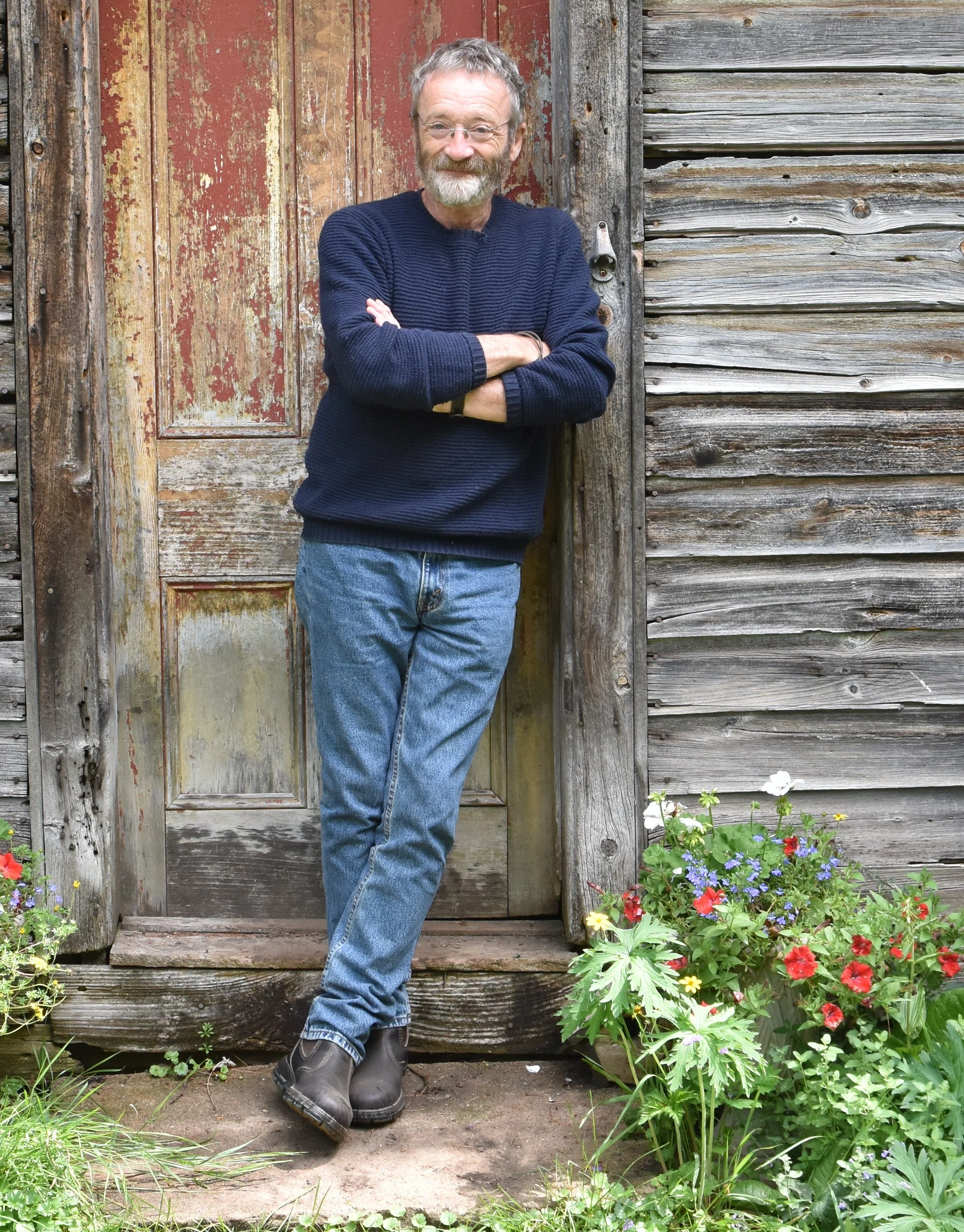 David Francey leans against a rustic door frame, arms crossed with a gentle grin