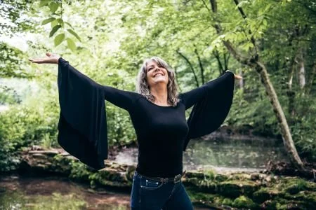Kathy Mattea stands with arms open, looking up and smiling by a creek
