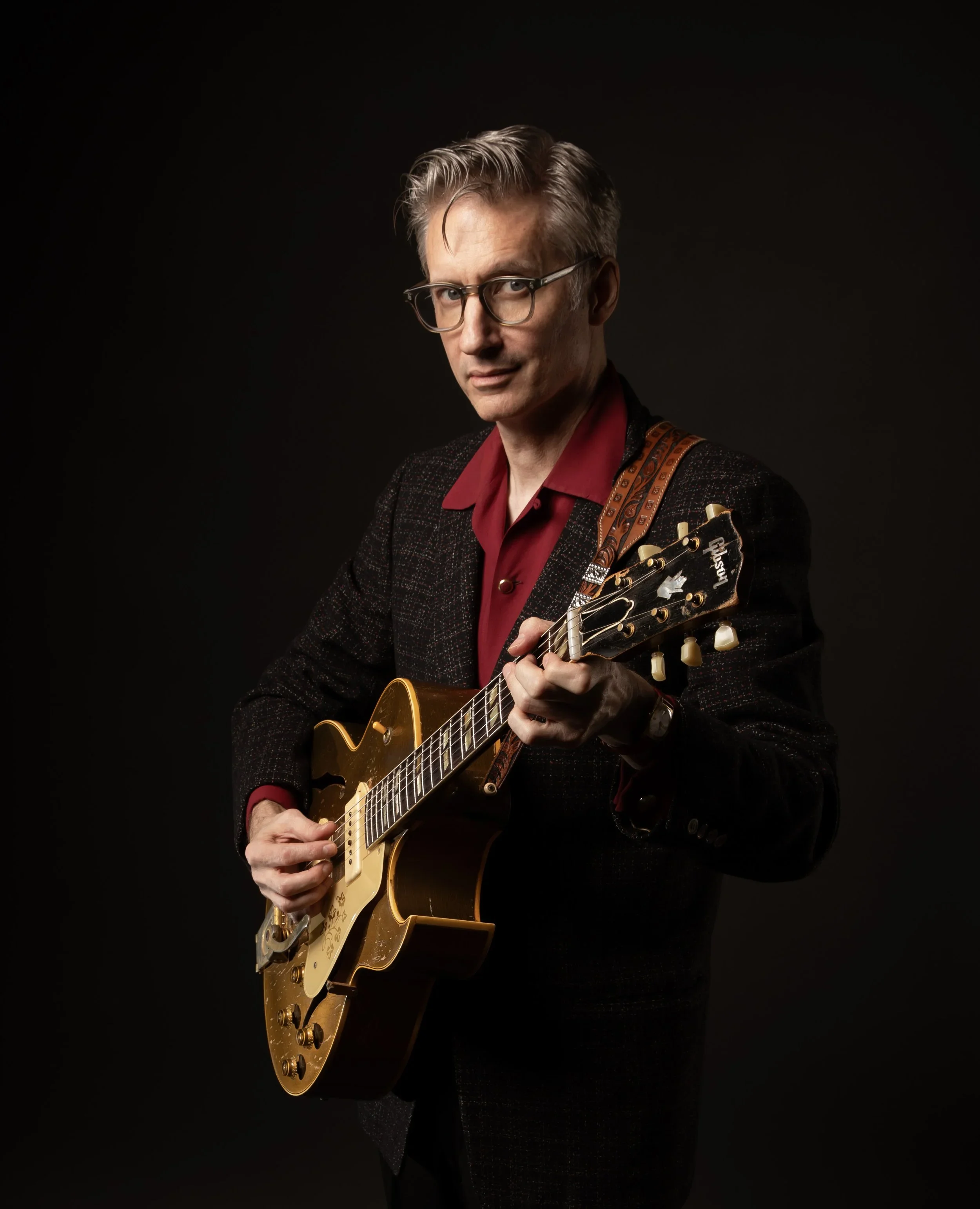 Joel Paterson holds a hollow-body electric guitar against a dark background, wearing glasses, a red shirt, and a dark jacket.