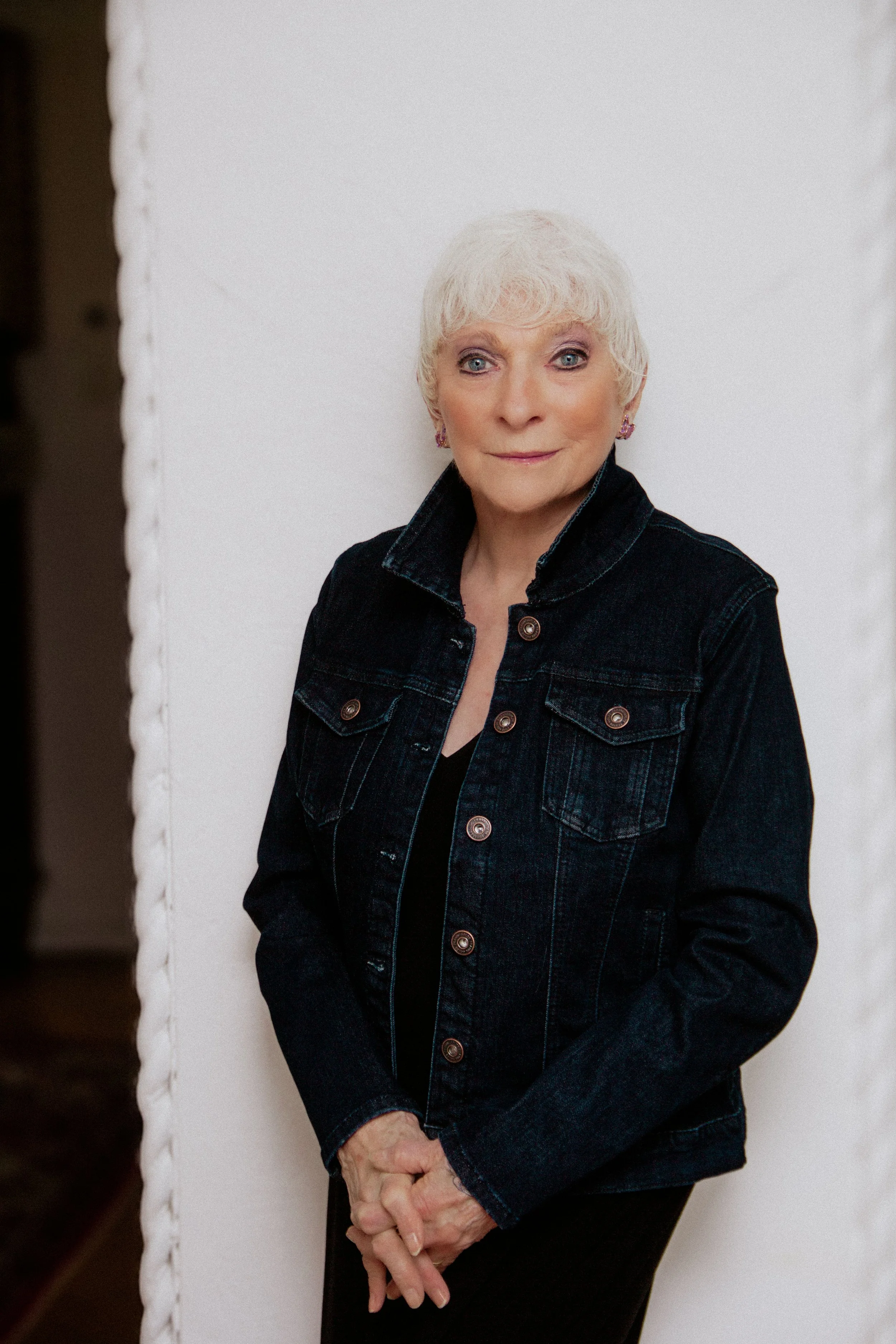 Judy Collins wears a denim jacket and stands before a white background with hands clasped