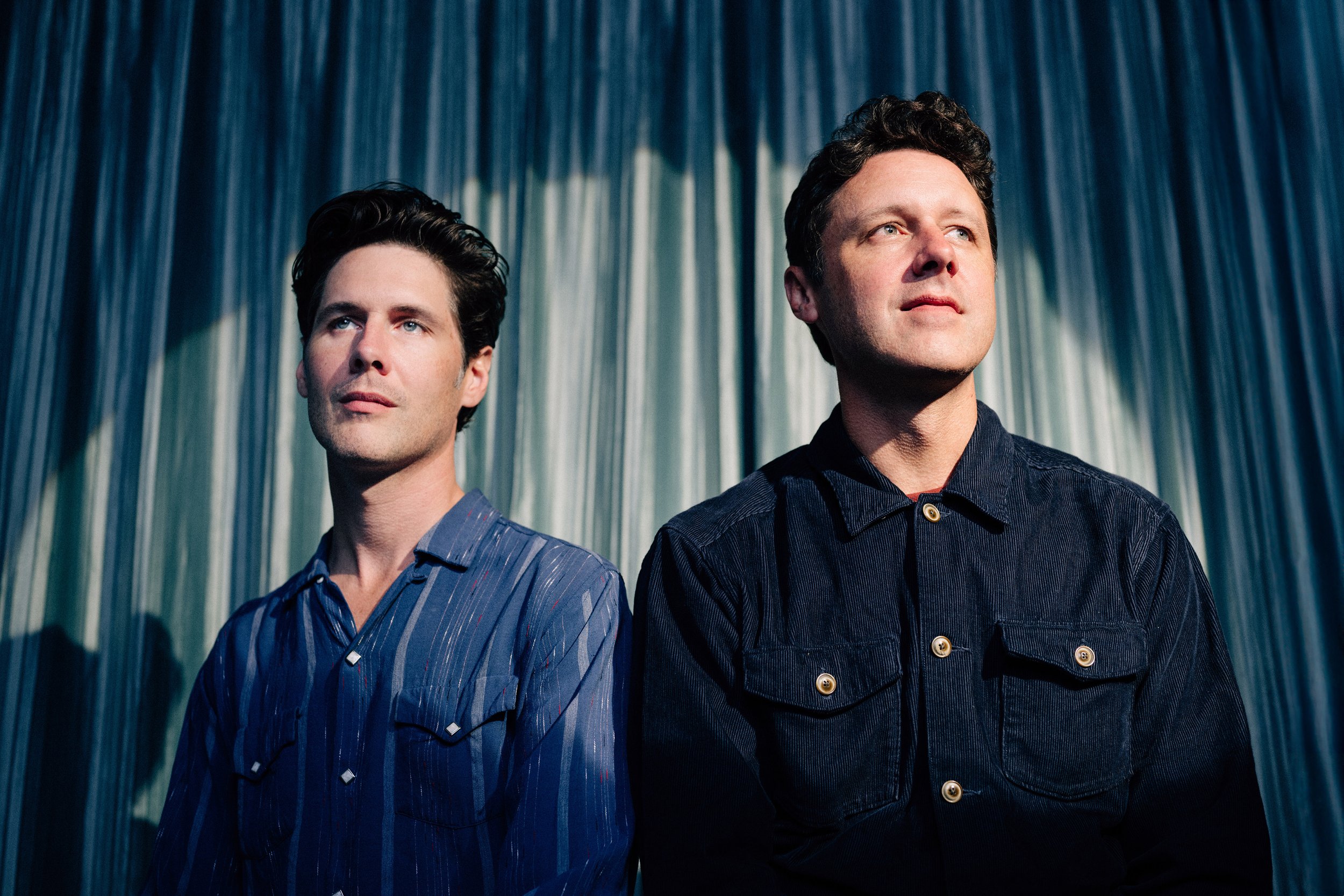 The Cactus Blossoms duo pose in front of a blue-and-white draped backdrop, both looking upward and away from the camera. One wears a blue striped shirt and the other a dark blue jacket.
