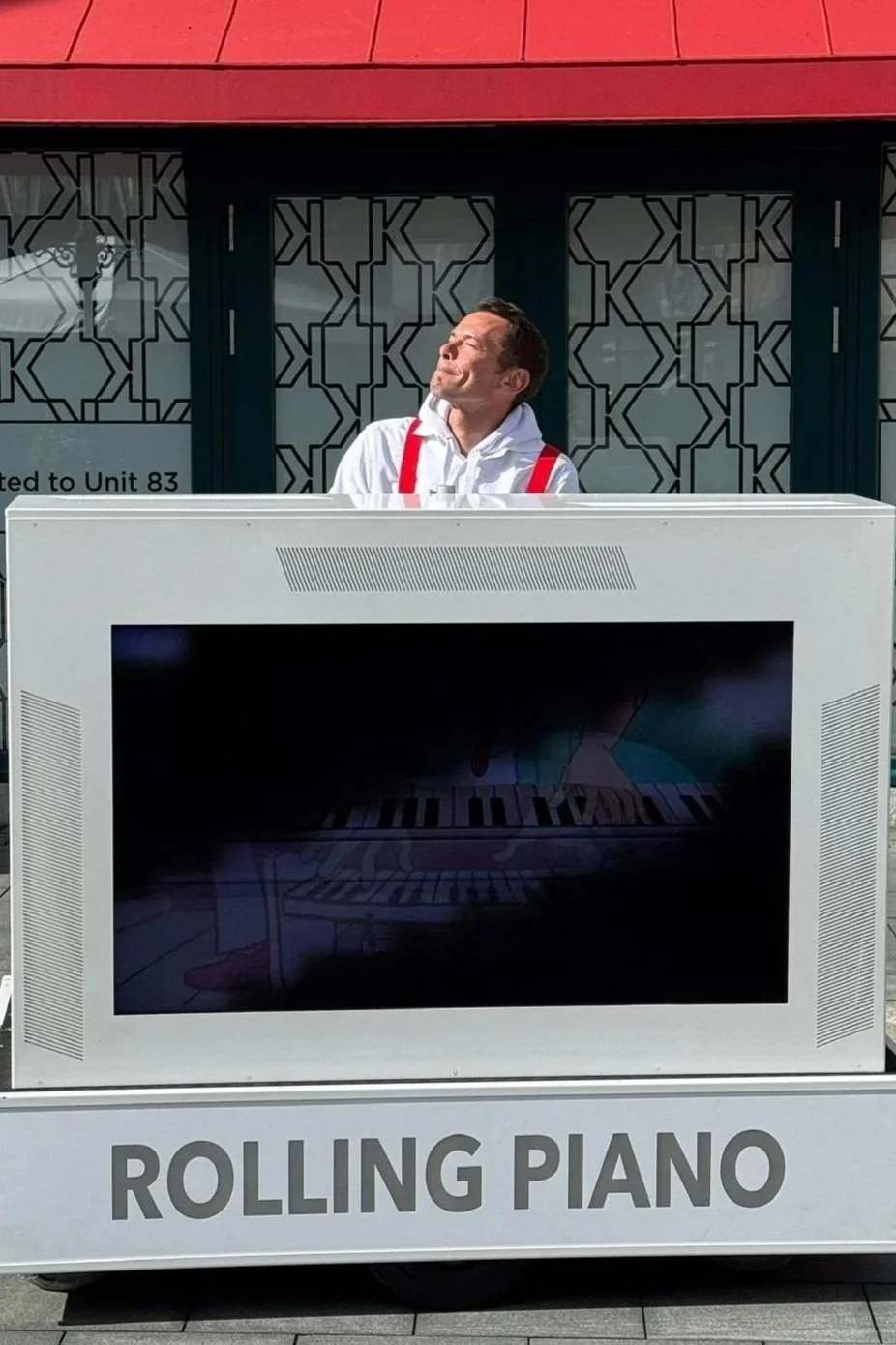Ein Mann steht hinter einem tragbaren Rollpiano mit der Beschriftung "ROLLING PIANO" vor einem Gebäude mit schwarzen Mustertüren und einem roten Dach. Der Mann trägt einen weißen Kapuzenpullover mit roten Suspenders und sonnt sich in der Sonne.