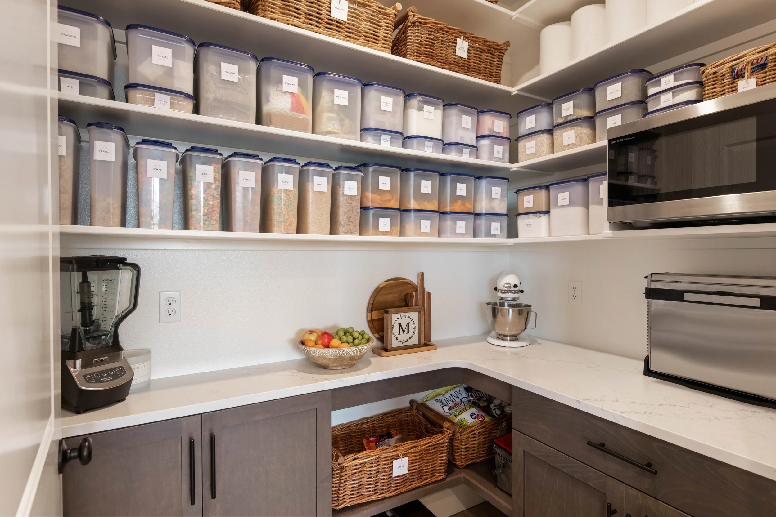 Pantry shelves filled with labeled clear plastic containers of dry goods like cereal, popcorn, crackers, flour, and sugar, along with woven baskets, a fruit bowl, a stand mixer, and a microwave oven.