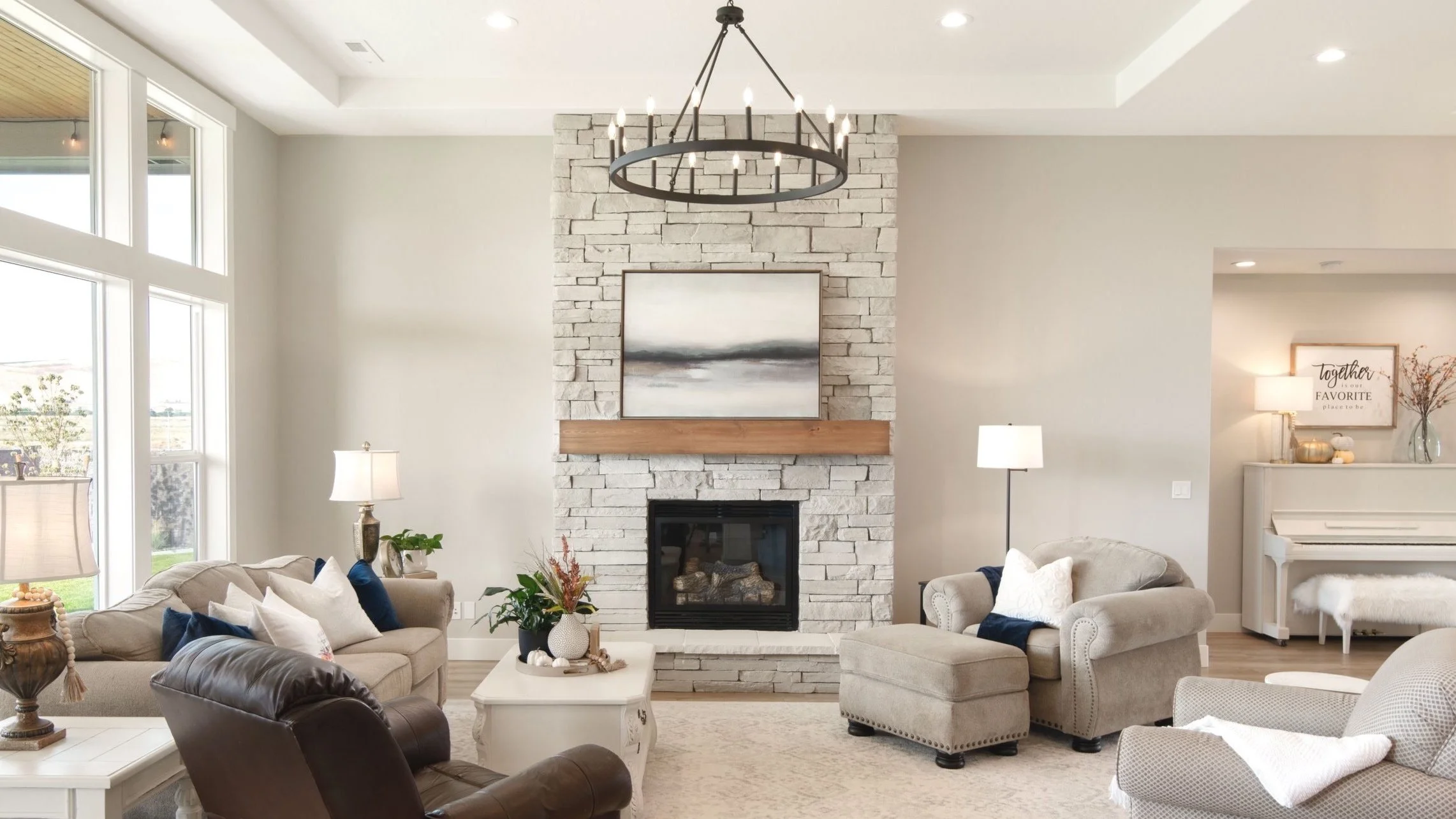 Living room with a white stone fireplace, circular chandelier, large windows, beige sofas, armchair, coffee table, lamps, and wall art.