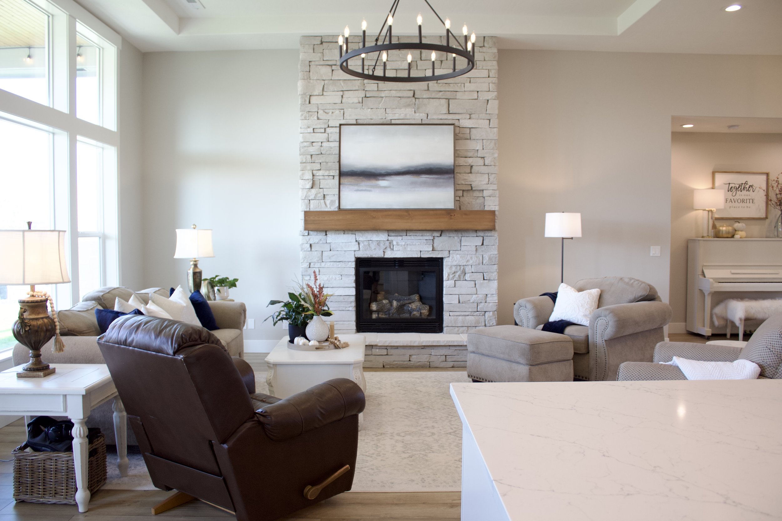 Living room with a white stone fireplace, a round chandelier, beige sofas, a leather armchair, and lamps, with large windows on the left and wall art on the right.