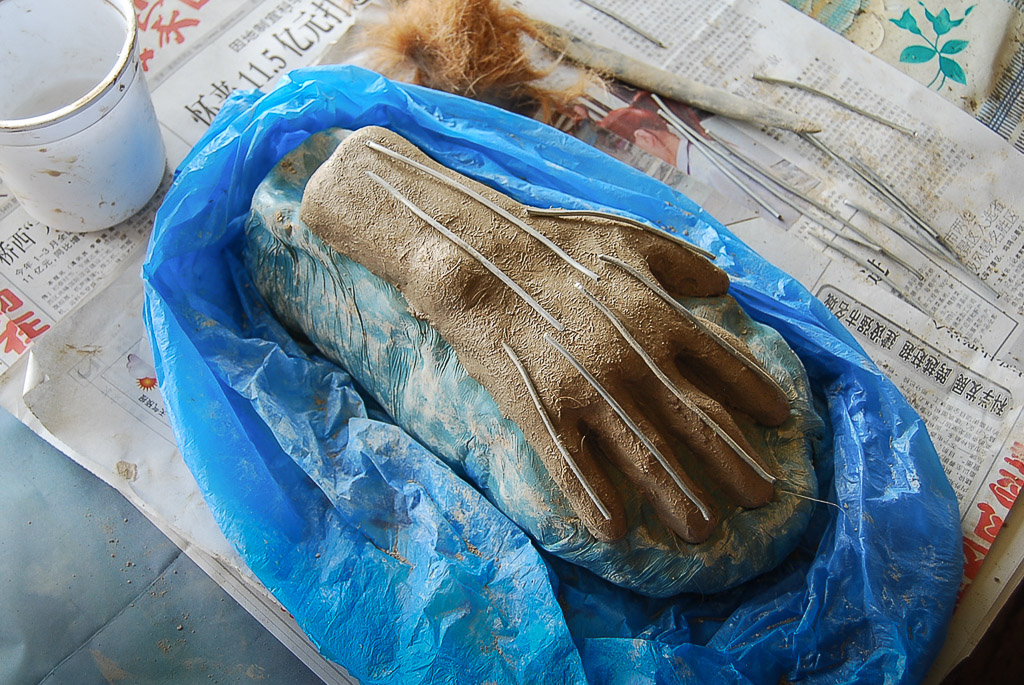 While the hand is still wet, metal pins are inserted as armature to further reinforce cohesiveness and structure. Here they are laid on top to gauge the right length. Behind the hand is a wad of reddish cow hair. 