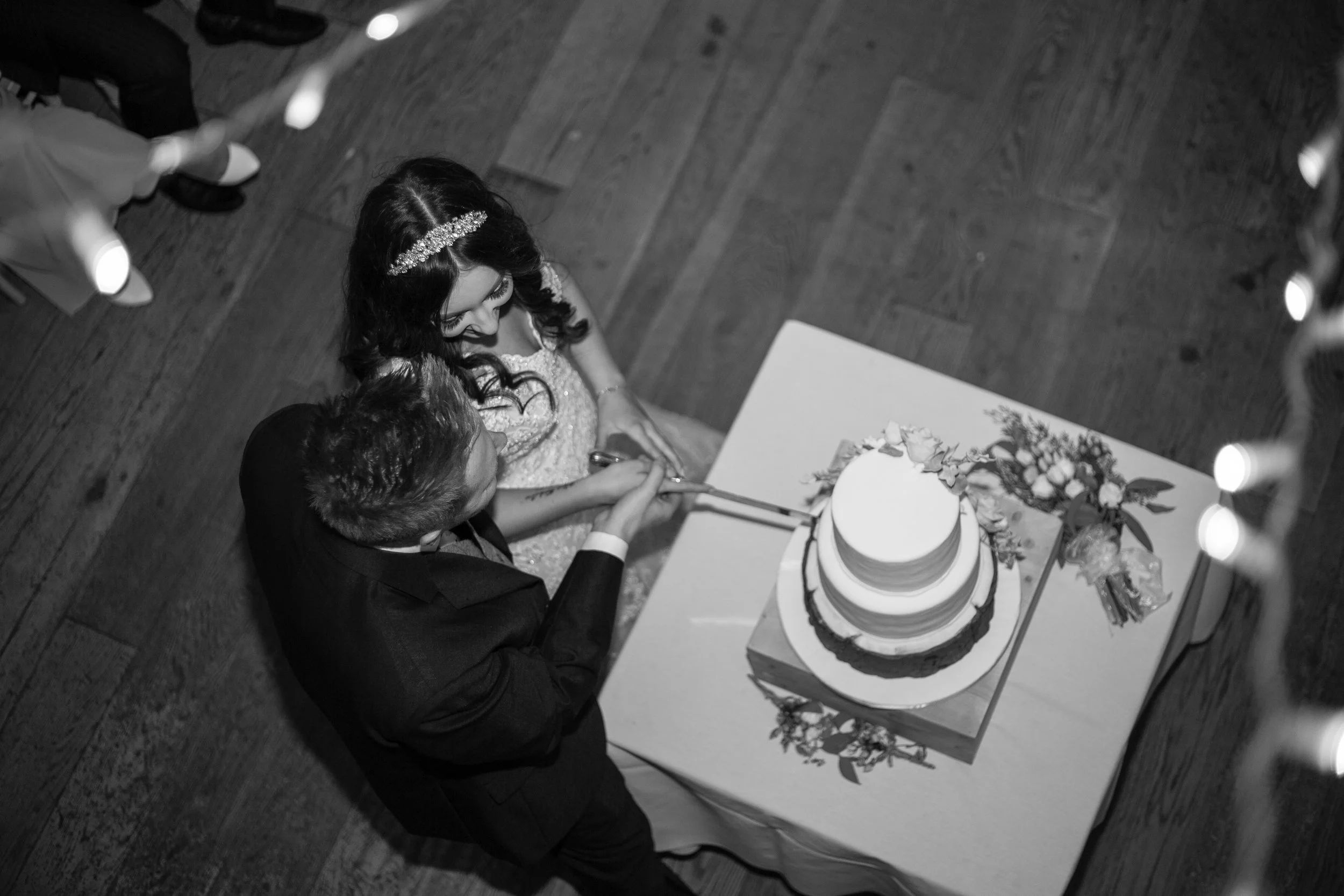 A bride and groom slicing a wedding cake at their reception, with a wedding bouquet on the table and guests in the background.