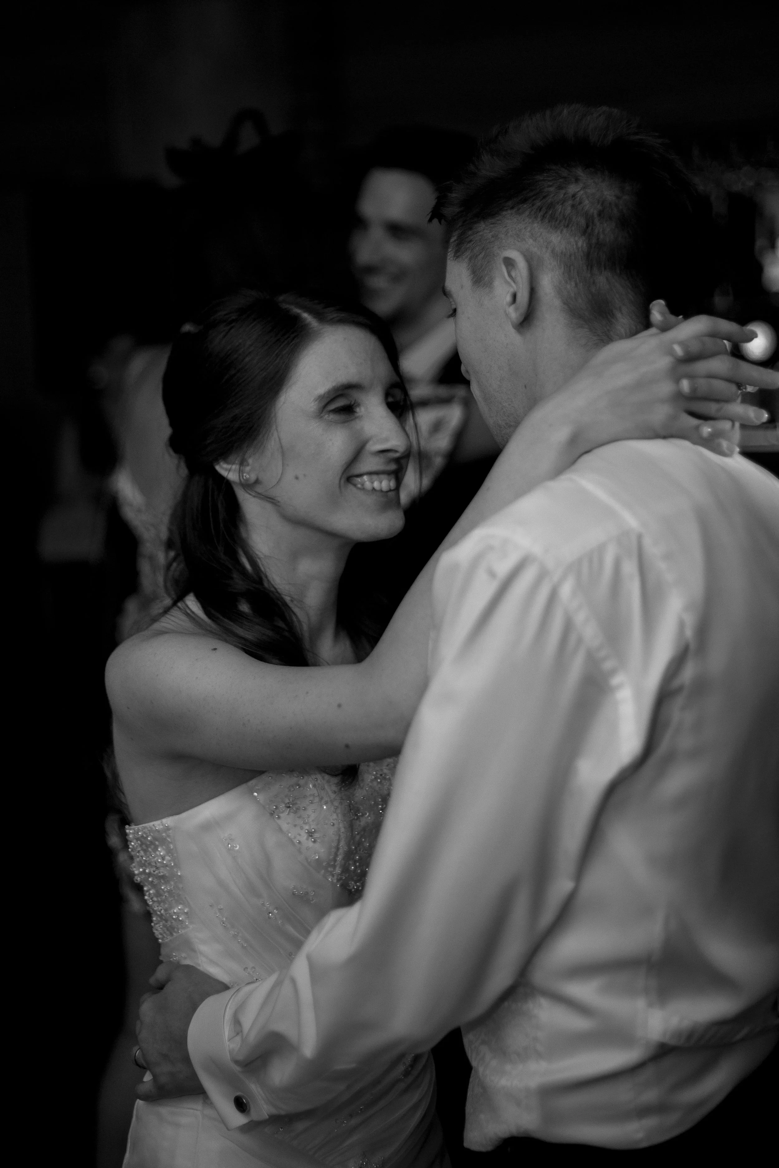 A black-and-white photo of a woman and a man dancing closely, smiling at each other. The woman has long dark hair and is wearing a strapless dress with embellishments. The man has short hair styled upward and is wearing a light-colored dress shirt. I