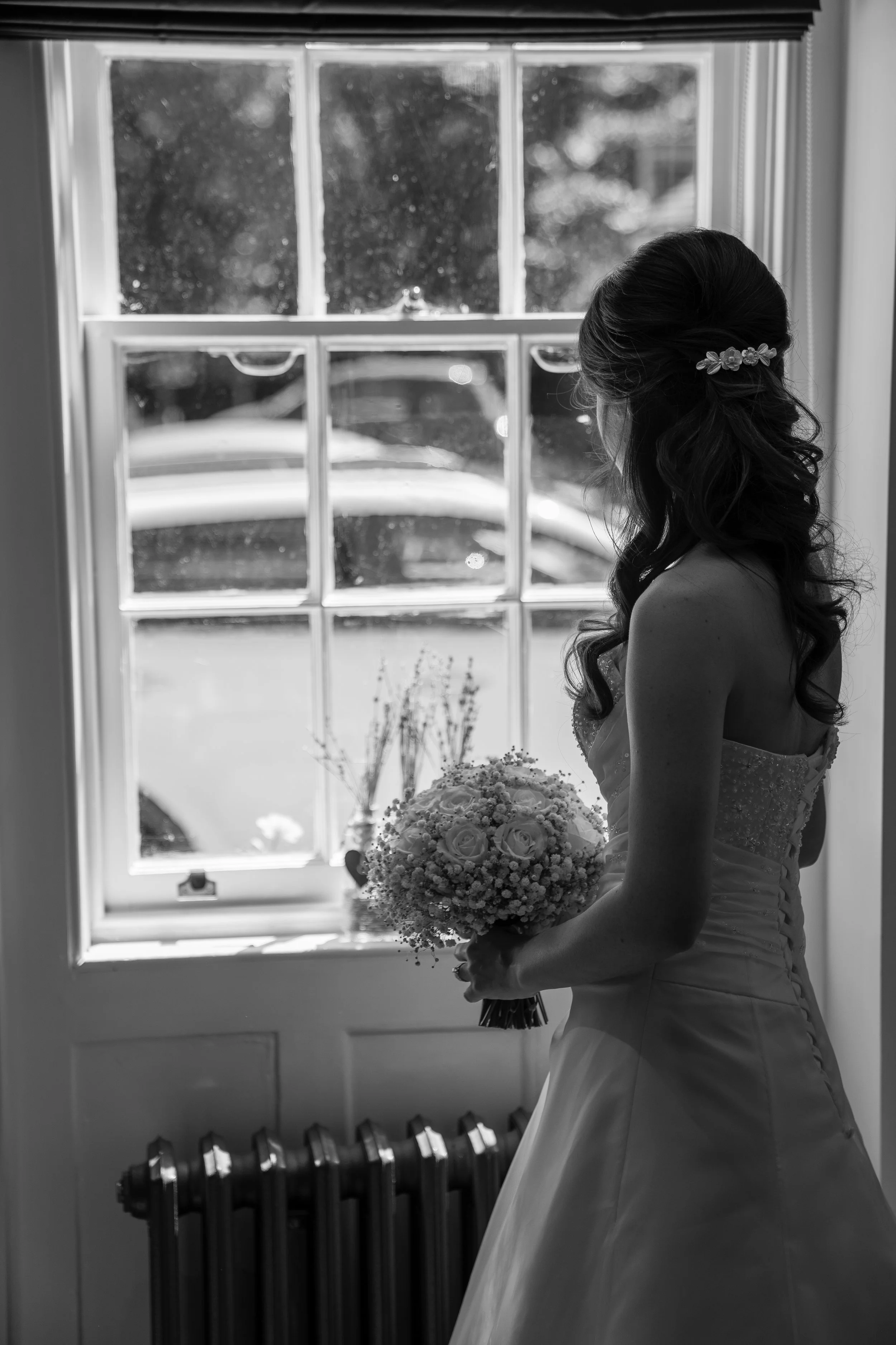 A bride in a wedding dress standing by a window, holding a bouquet of flowers, with her face turned away from the camera.