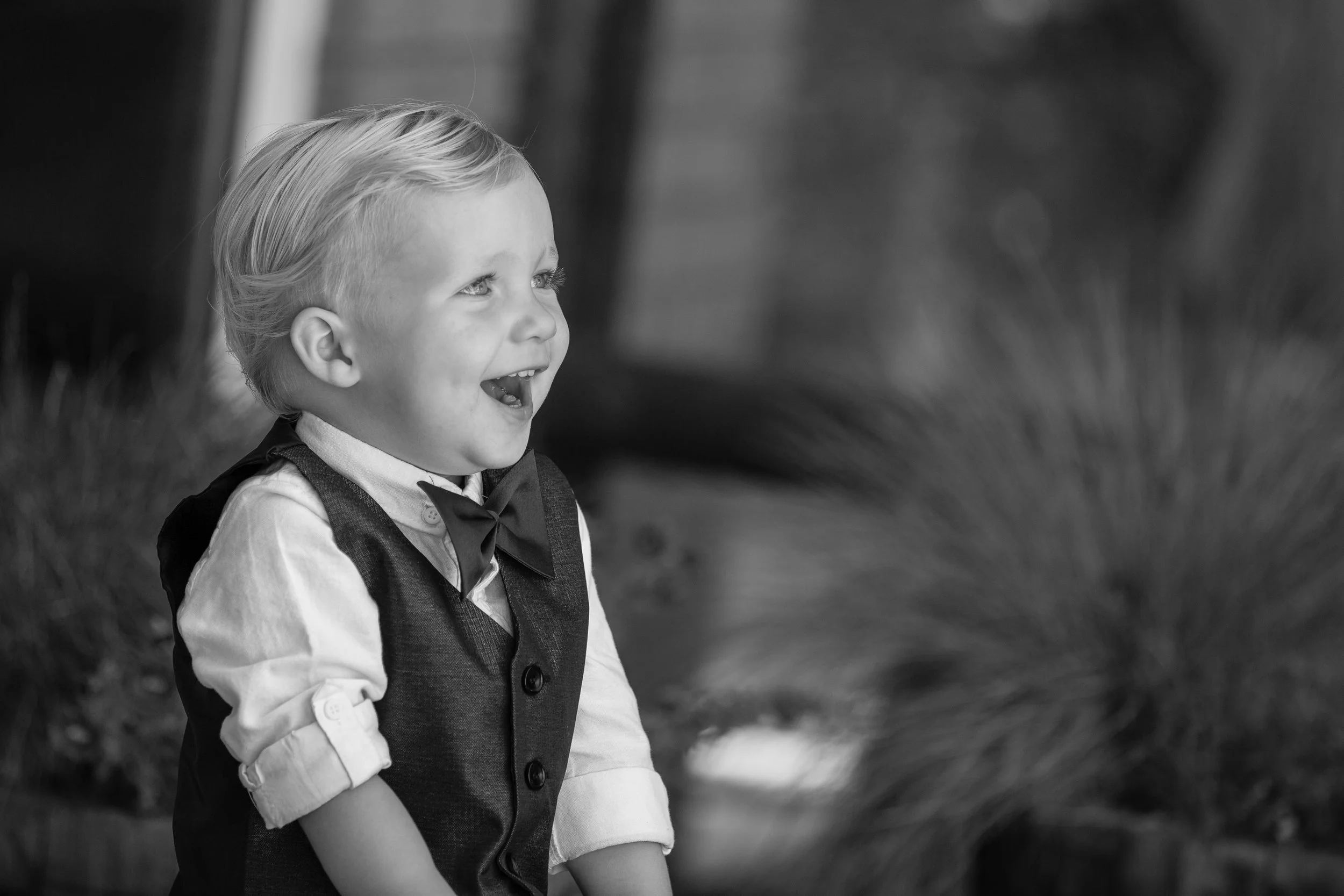 A young boy with light-colored hair, wearing a dress shirt, vest, and bow tie, is laughing or smiling outdoors.