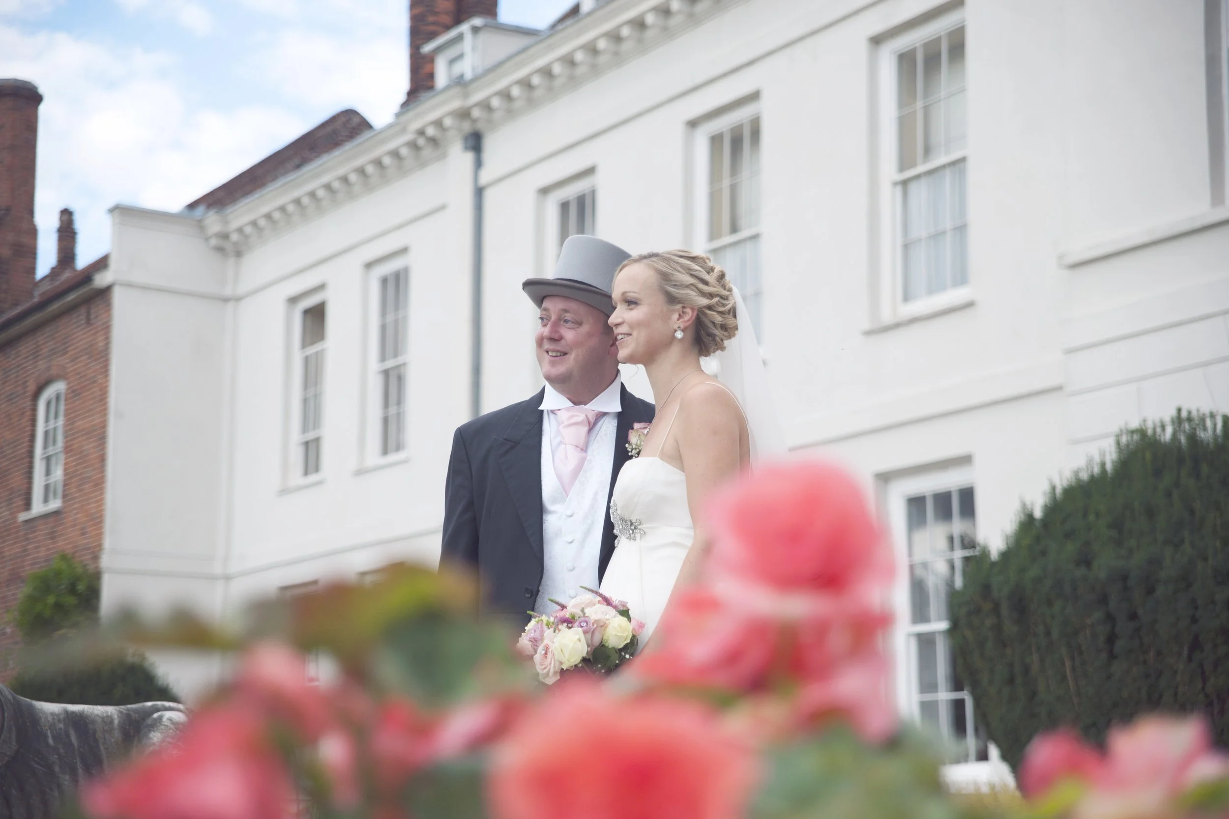 A bride and groom standing outdoors on their wedding day with a white building in the background. The bride is holding a bouquet of pink and white flowers. The groom is wearing a black suit with a pink tie and a gray top hat.