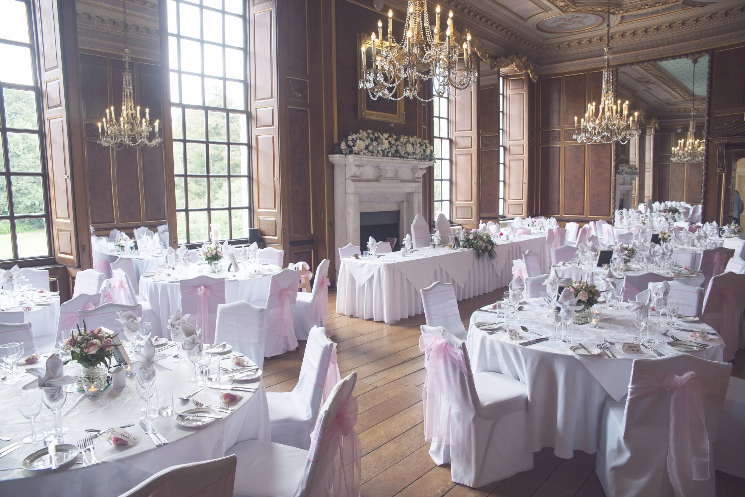 Elegant banquet hall decorated for a wedding reception with white tables, chairs with pink sashes, floral centerpieces, and chandeliers.