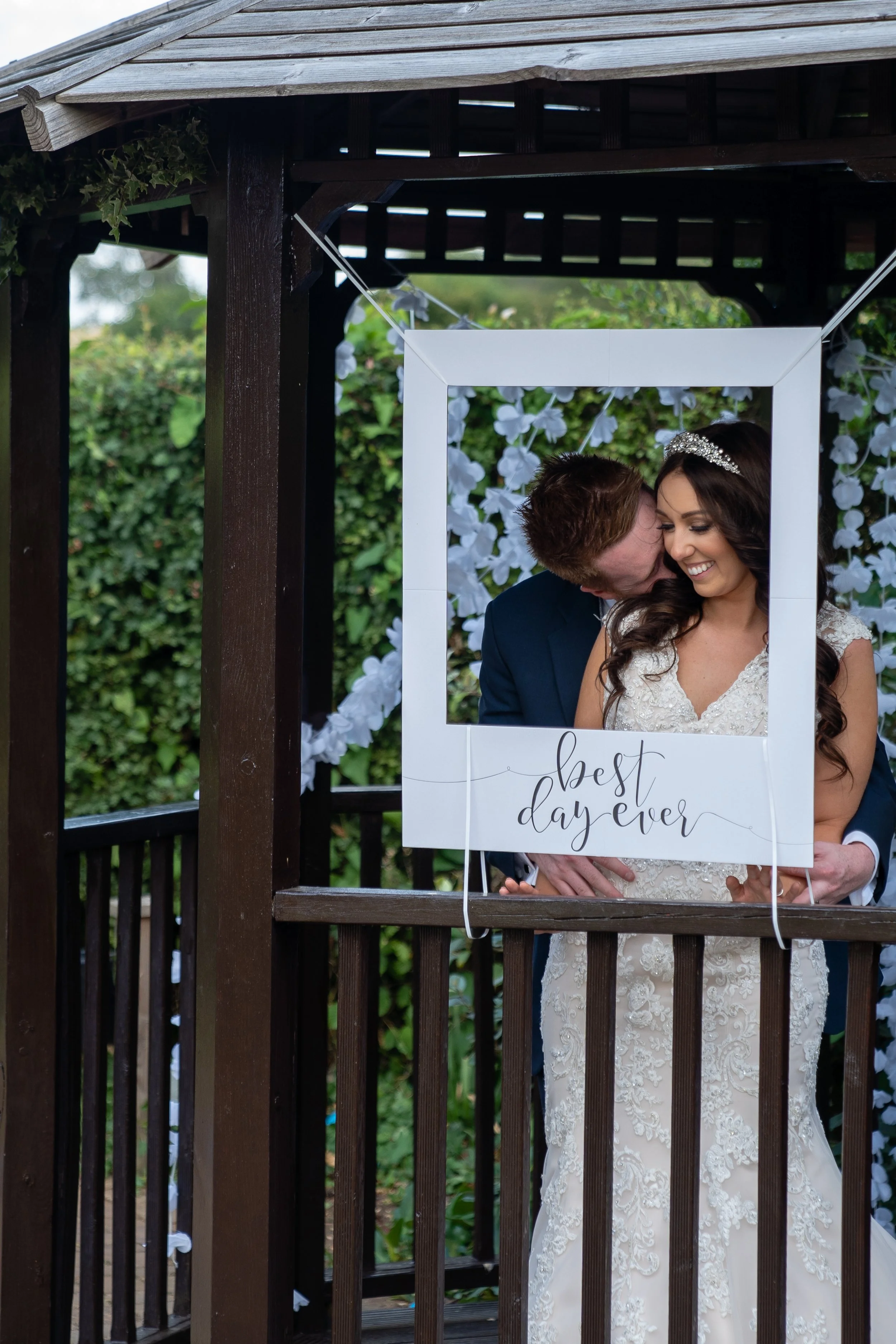 A bride and groom on a wooden balcony, with the groom kissing the bride on the cheek, during their wedding celebration. The bride is smiling and holding a frame that reads 'best day ever'.