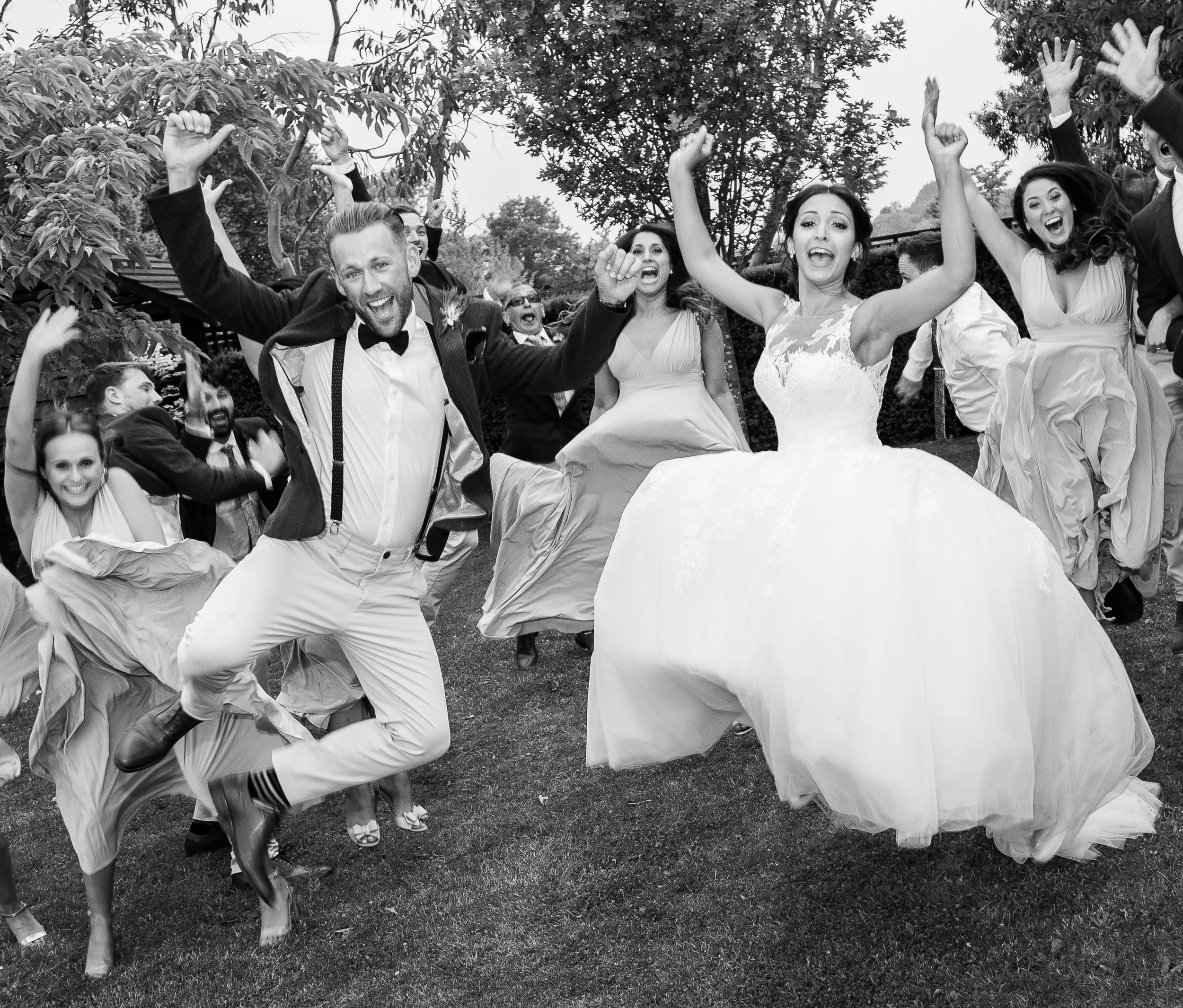 Black and white photo of a wedding celebration with joyful guests dancing and jumping outdoors.