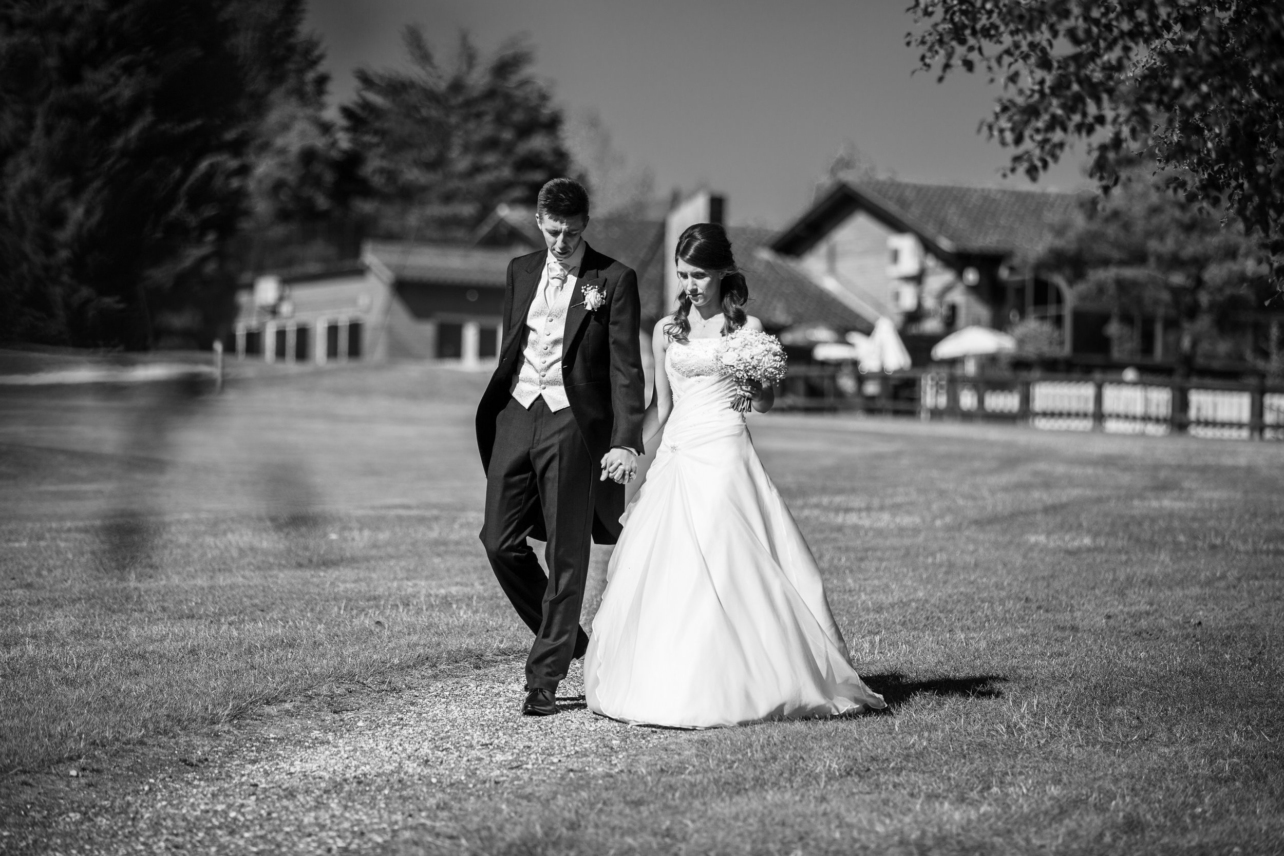 Black and white photo of a bride and groom holding hands and walking on a grassy path outdoors, with houses and trees in the background.