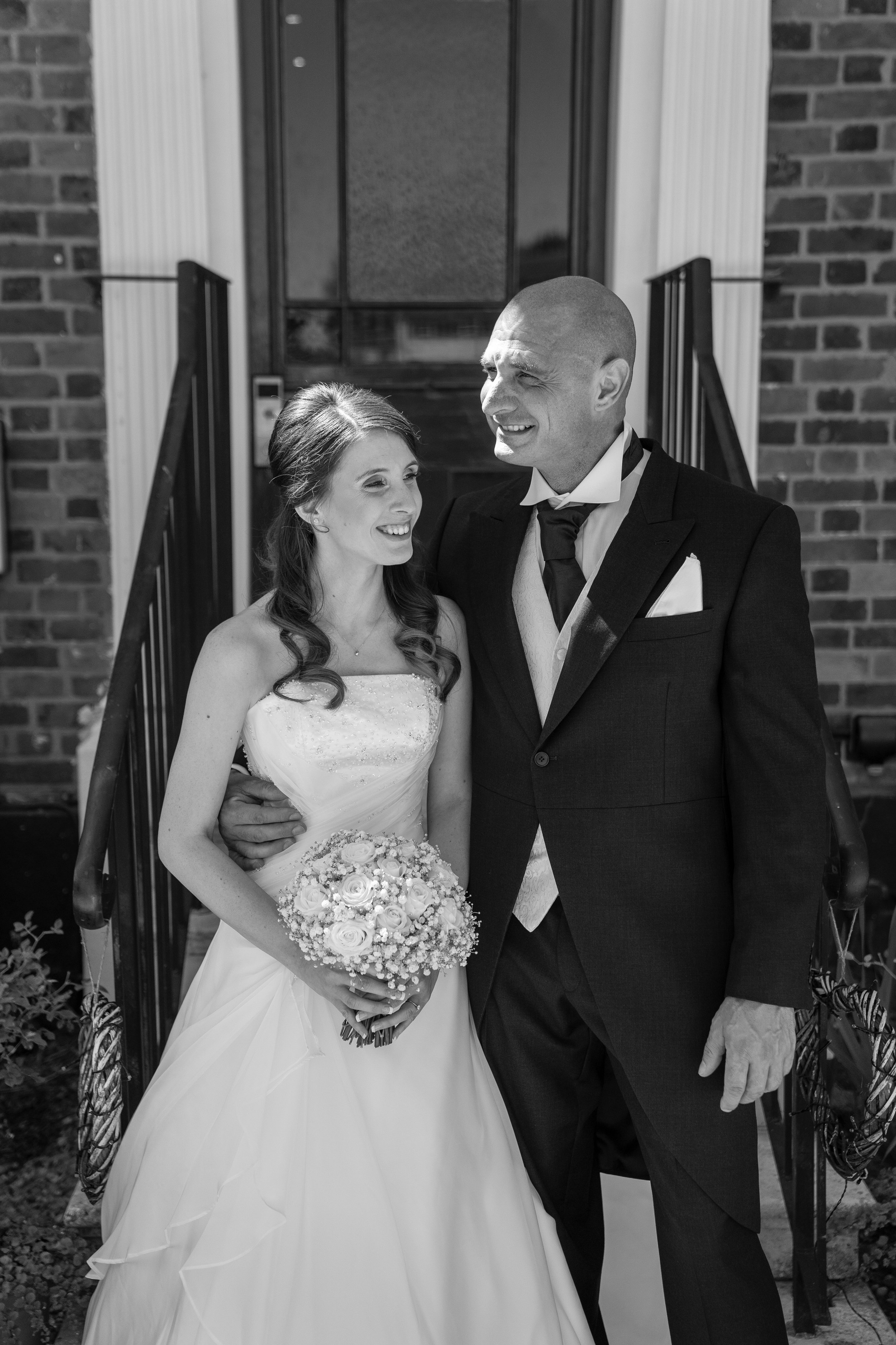 A bride in a wedding gown holding a bouquet of flowers, standing next to a man in a suit outside a house entrance.