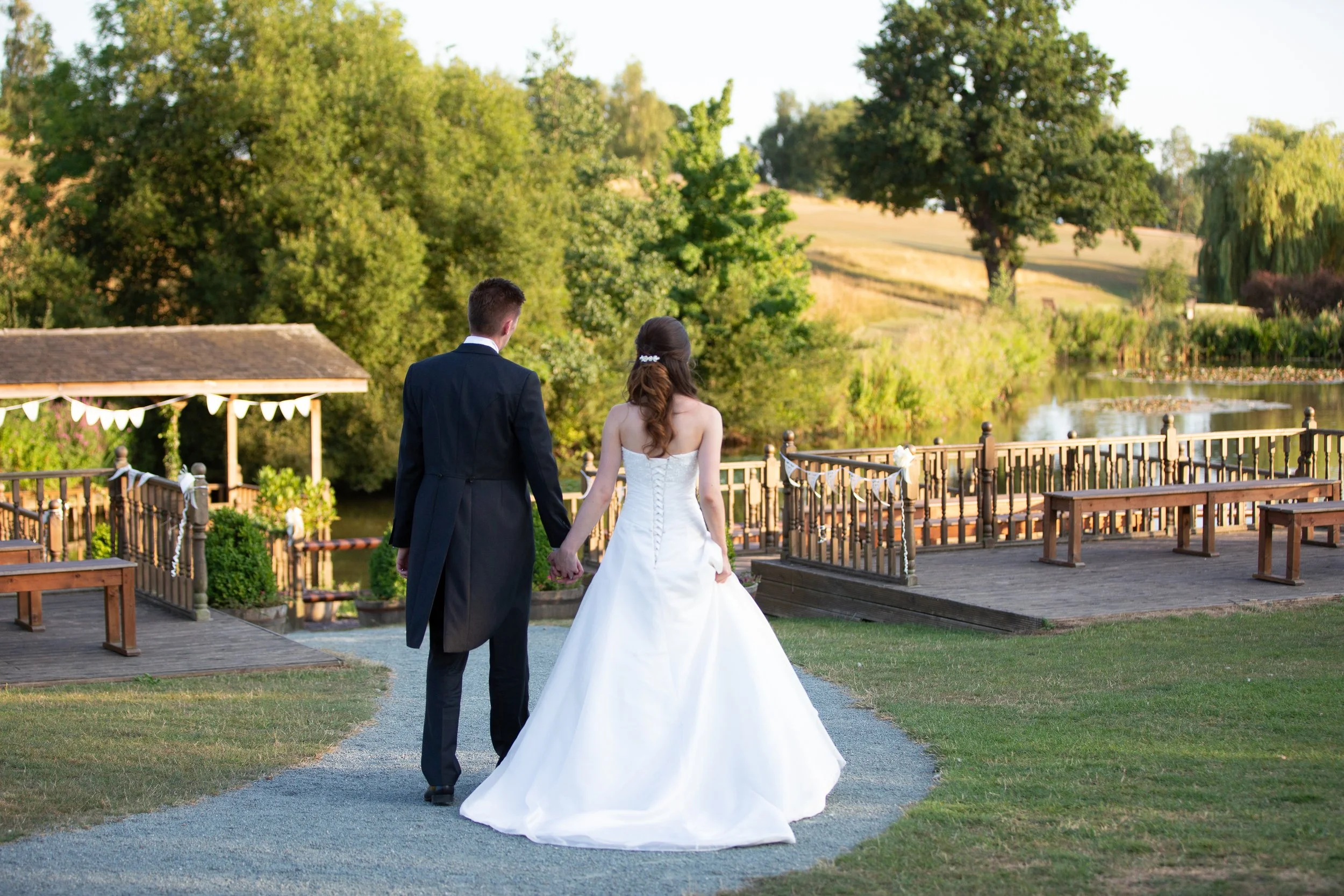 Bride and groom walking hand in hand on a pathway near a lake during sunset, with trees and a wooden gazebo decorated with bunting in the background.