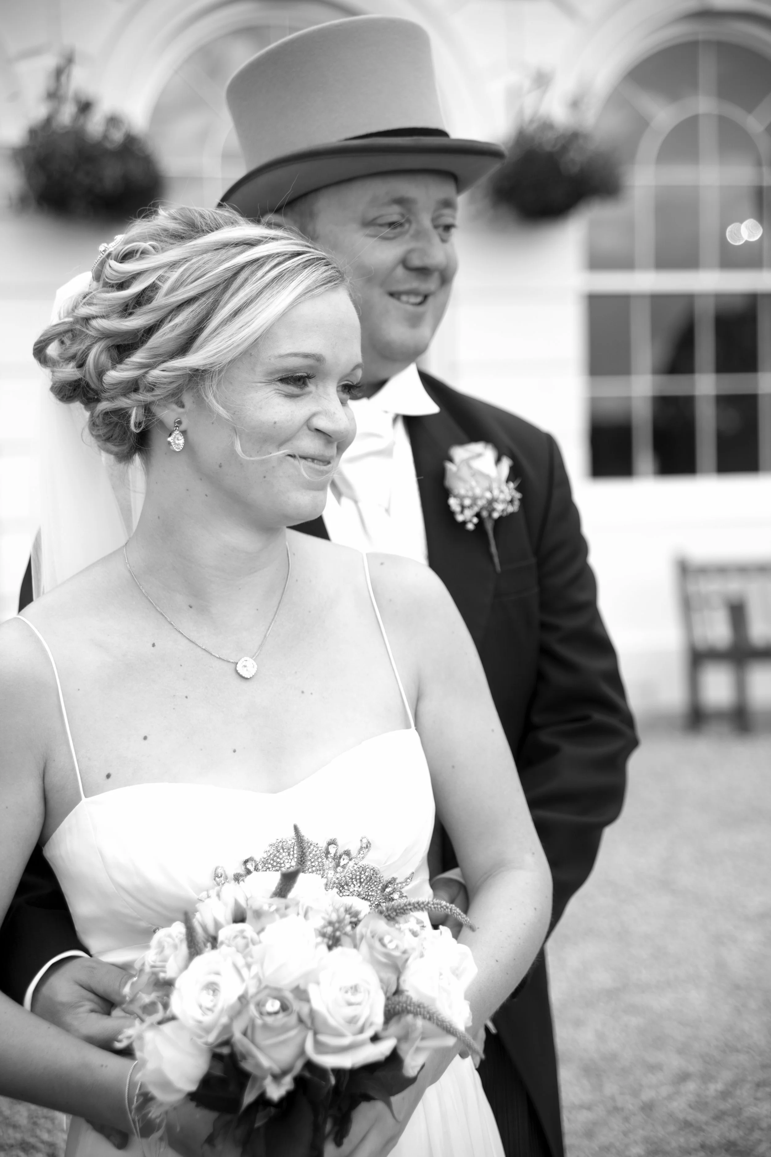 A bride and groom at their wedding, with the bride holding a bouquet of roses, dressed in a wedding gown and jewelry, and the groom wearing a suit with a top hat and boutonniere, standing outdoors with a building and window in the background.