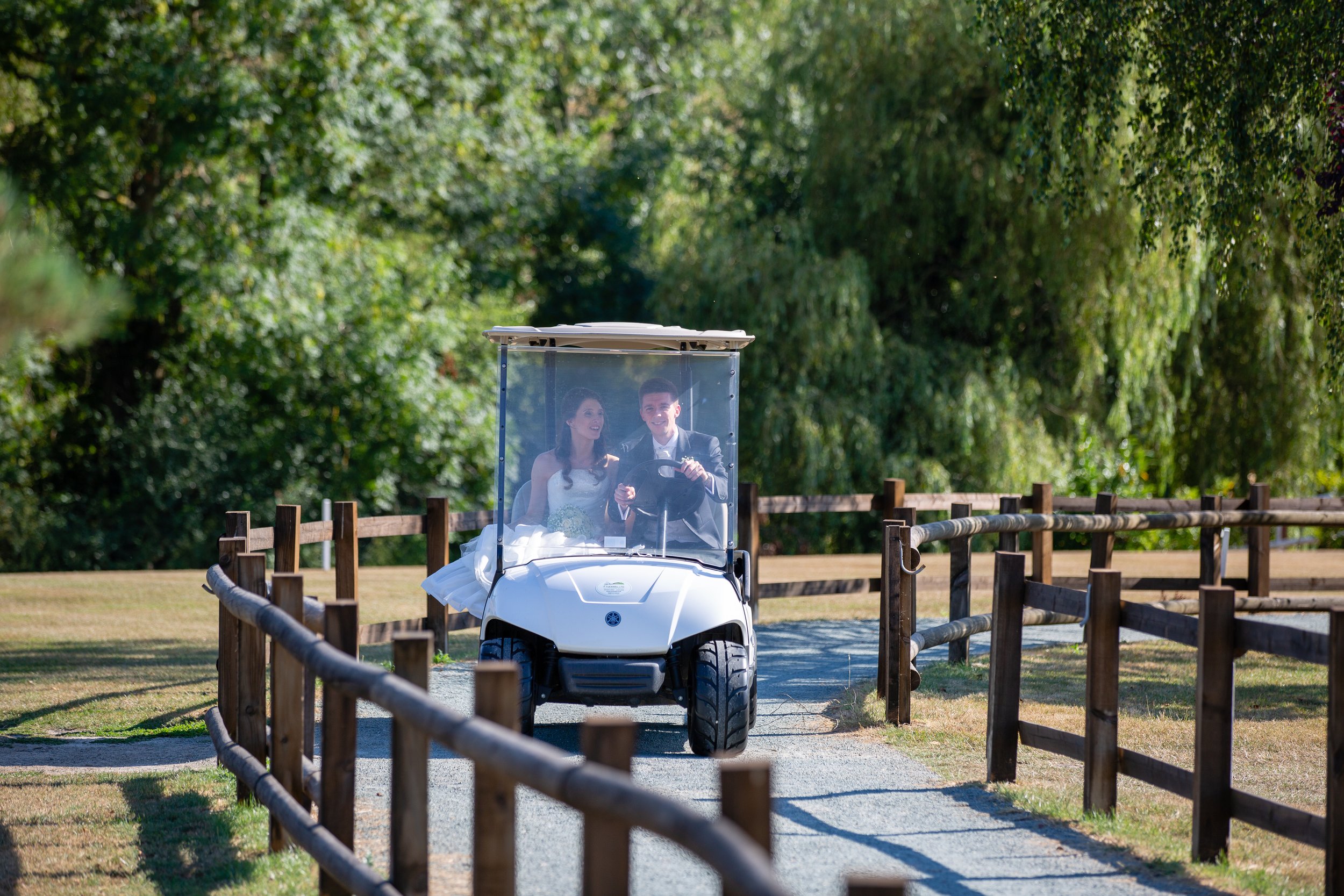 A bride and groom riding in a golf cart along a gravel path bordered by wooden fences, surrounded by lush green trees.
