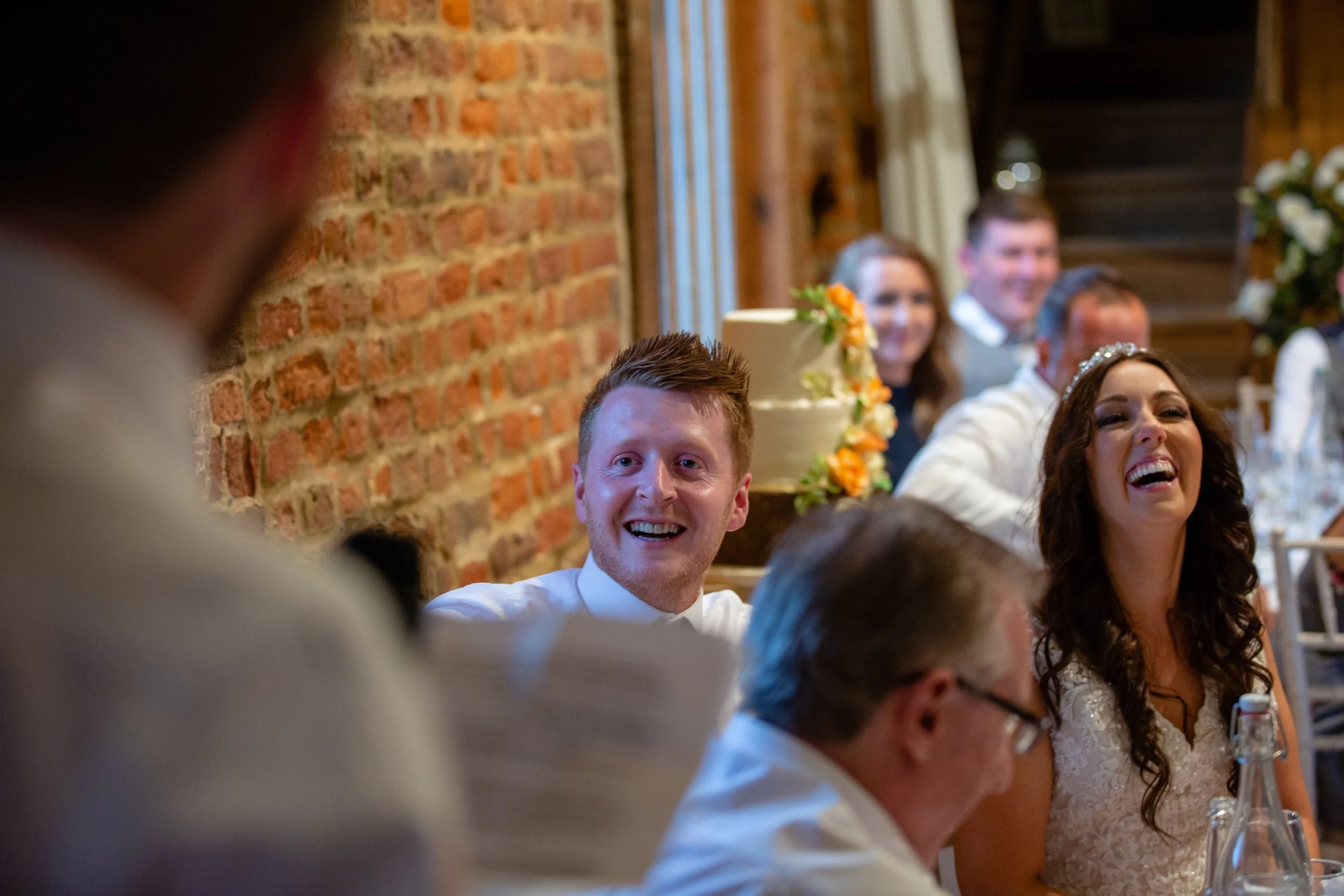 People sitting at a wedding reception table, smiling and laughing, with a wedding cake decorated with flowers in the background.