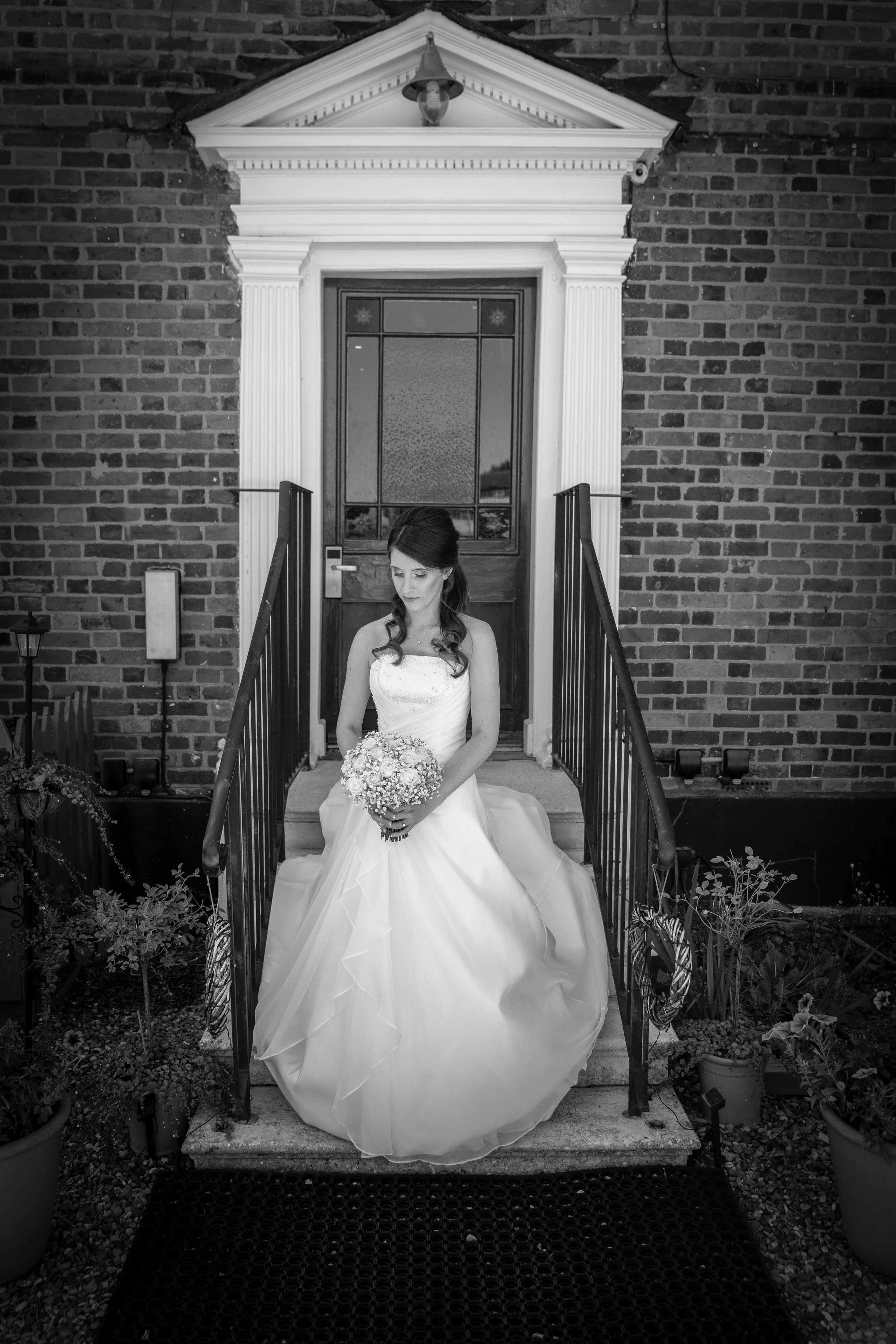 A bride in a white wedding gown sitting on the steps outside a brick house, holding a bouquet of flowers.