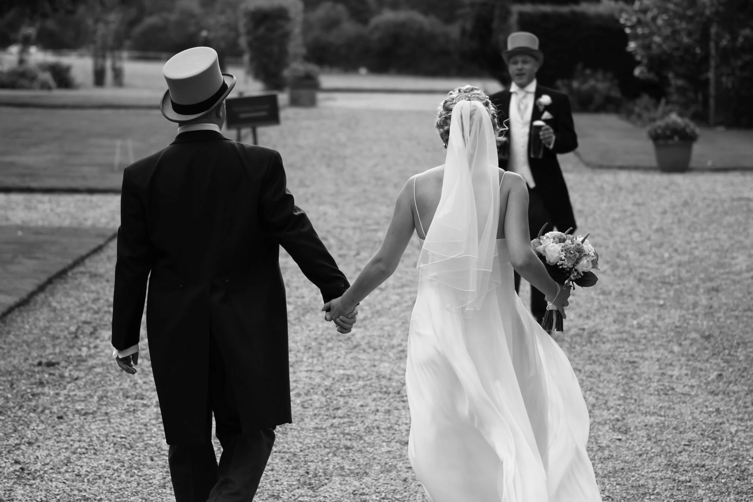 A bride and groom holding hands walking outside at their wedding, with a man in the background holding a drink, on a gravel path with trees and potted plants.