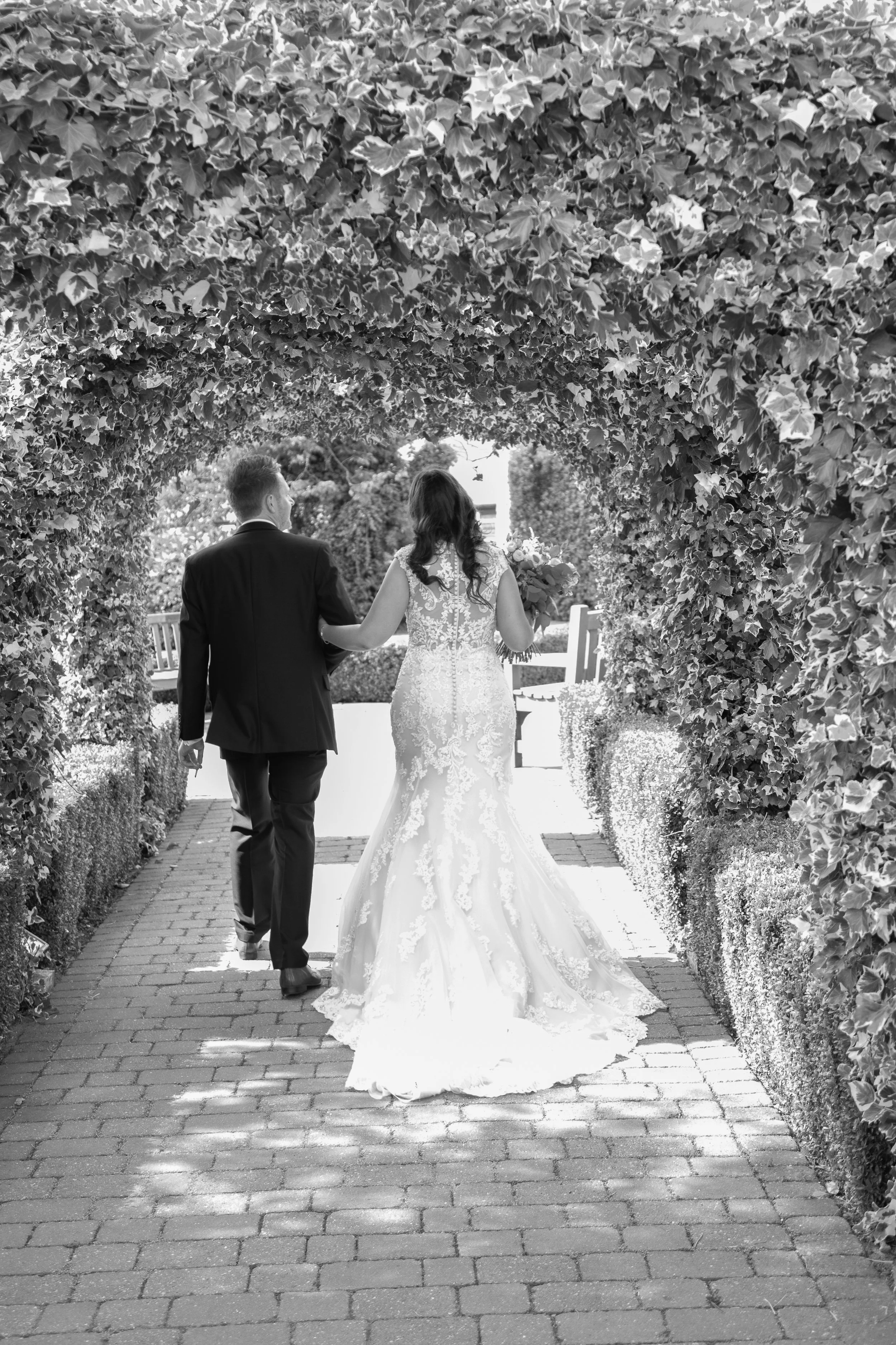 A bride and groom walking together under a leafy arch in black and white. The bride wears a lace wedding dress and holds a bouquet, while the groom wears a suit.