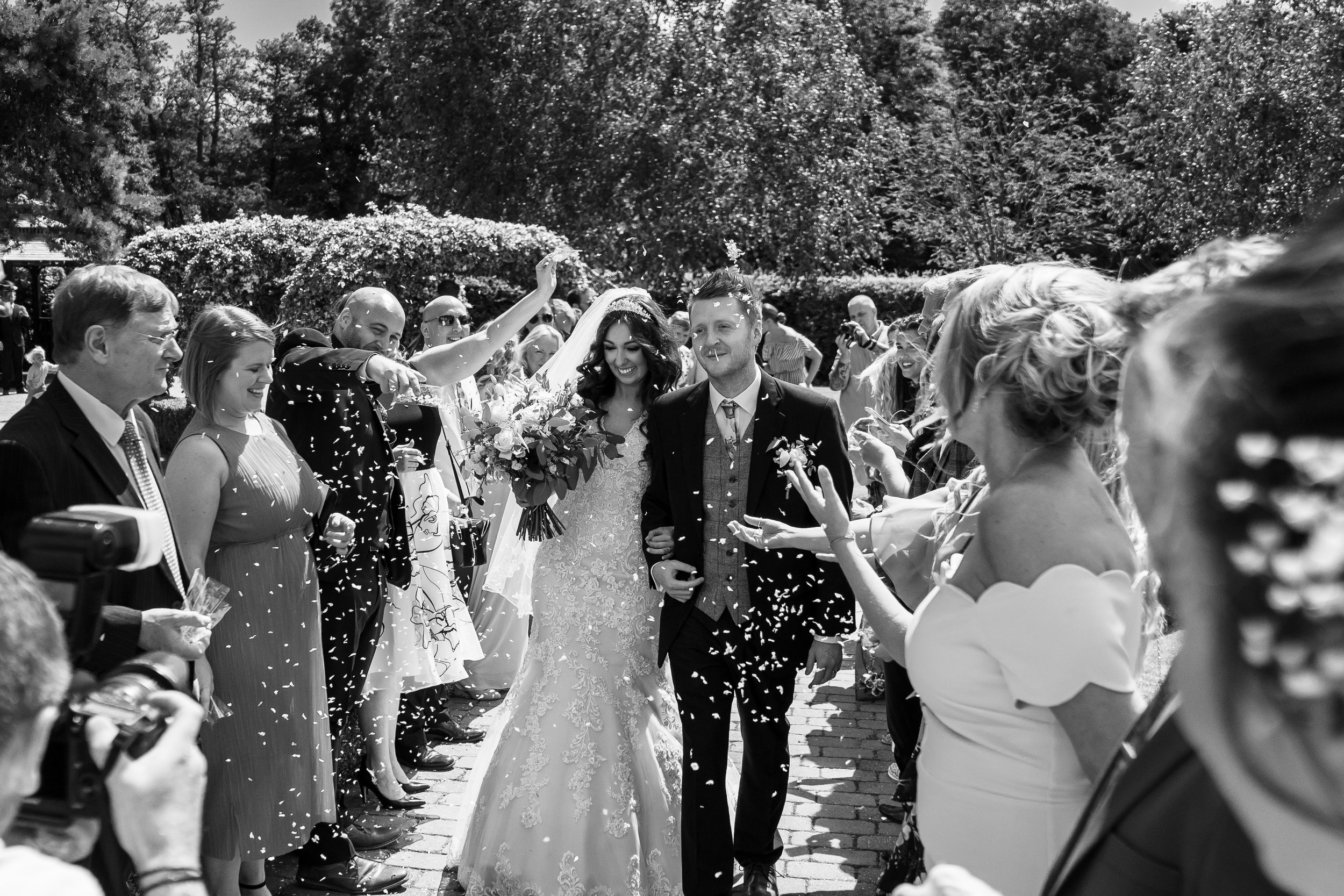 A black and white photo of a wedding celebration with a bride and groom walking down an aisle surrounded by friends and family, with confetti in the air.