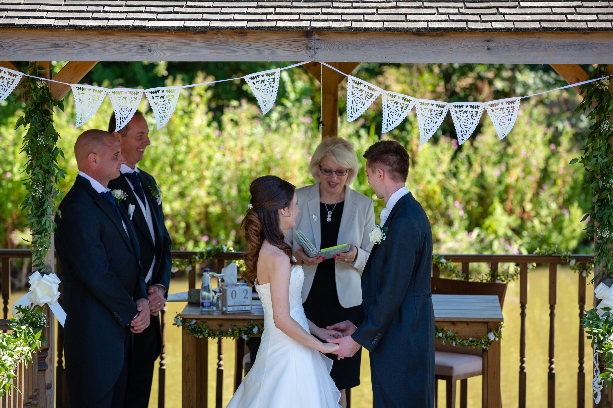 A couple getting married under a decorated wooden gazebo beside a pond, exchanging vows while an officiant oversees the ceremony, with three groomsmen watching. The scene is outdoors with lush greenery and decorative flags overhead.