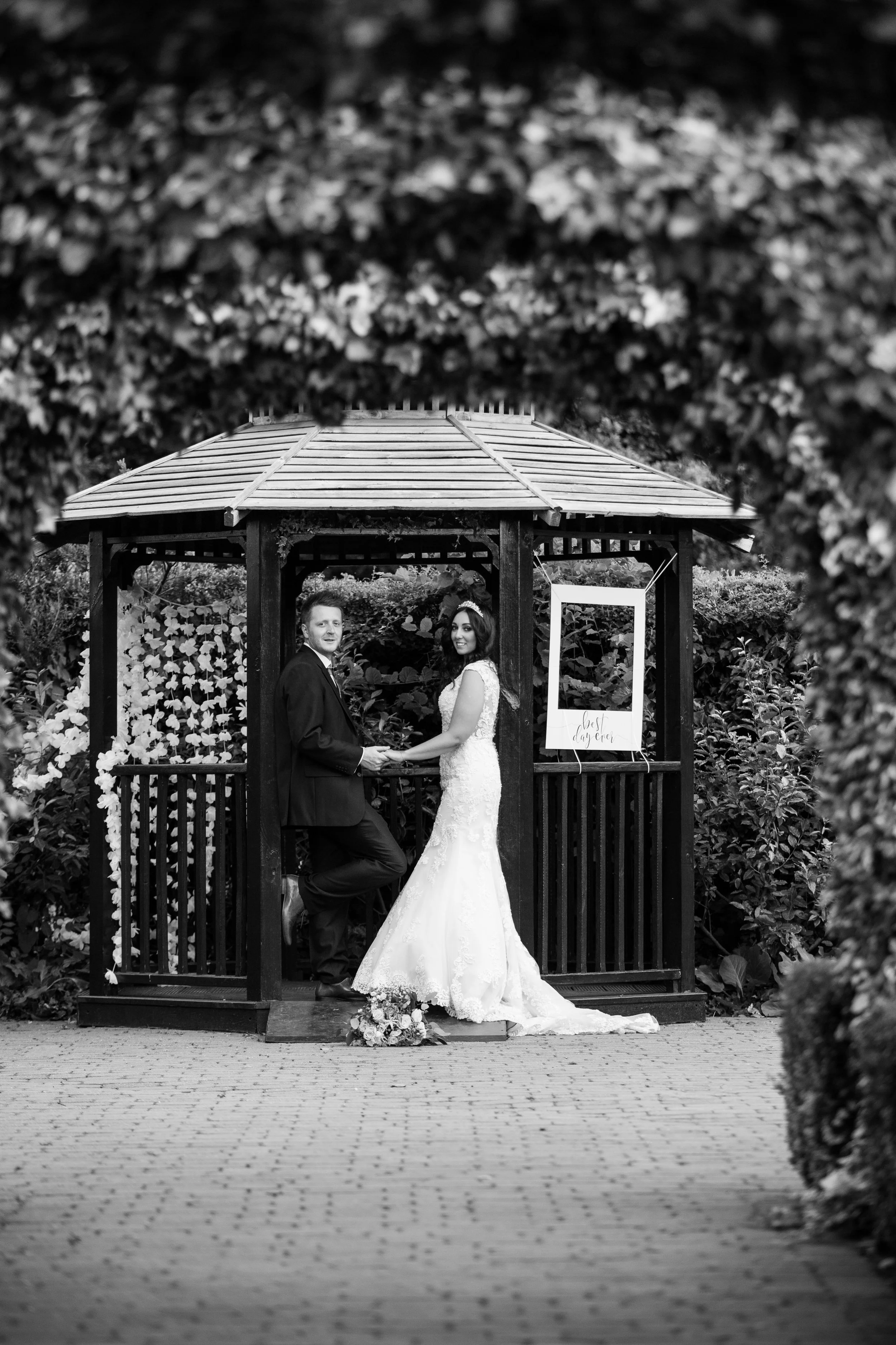 Black and white photo of a bride and groom holding hands inside a small wooden gazebo, surrounded by lush foliage, with a sign that says 'best day ever' on the side.