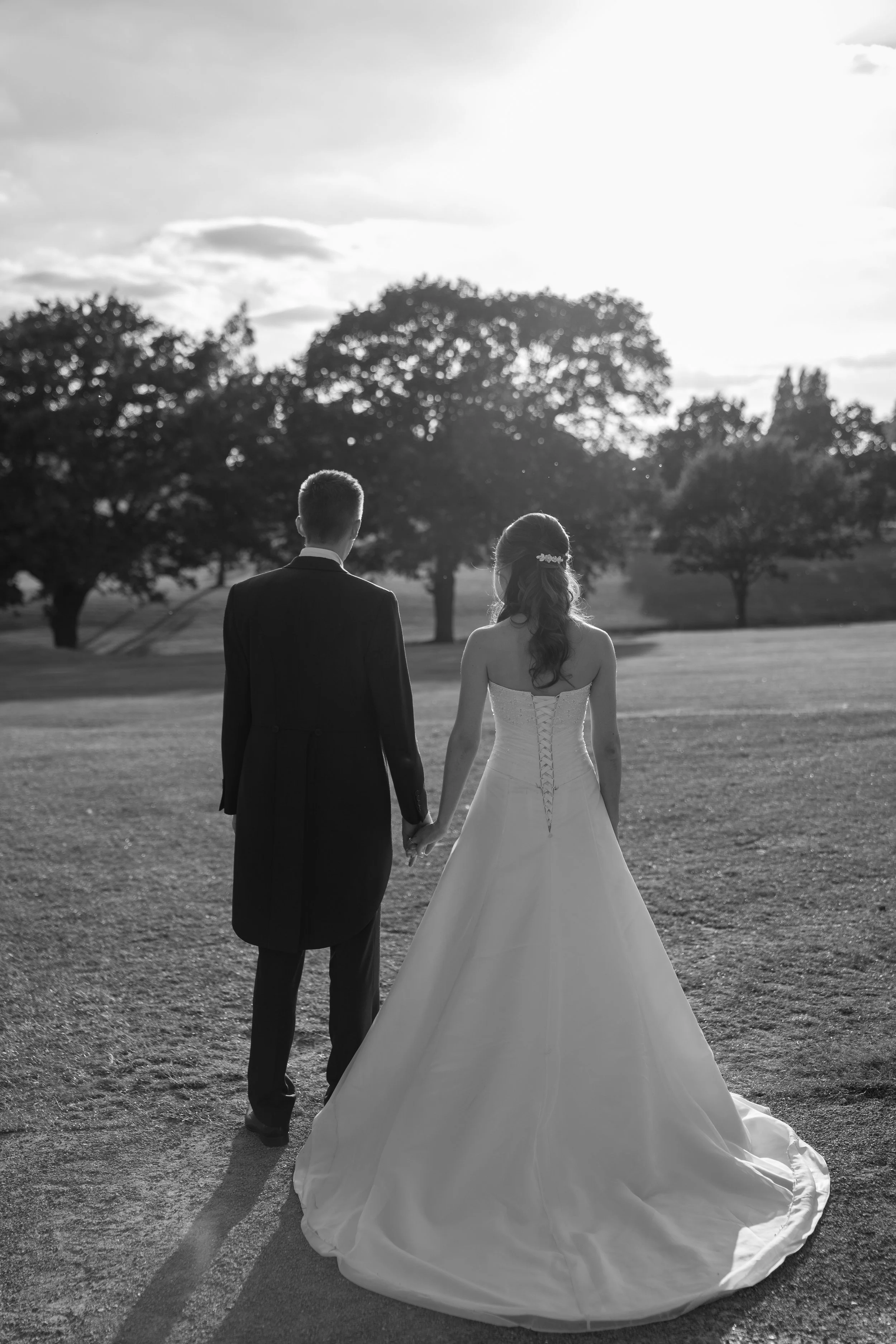 A black and white photograph of a bride and groom holding hands, walking outdoors on a grassy field with trees in the background, during sunset.