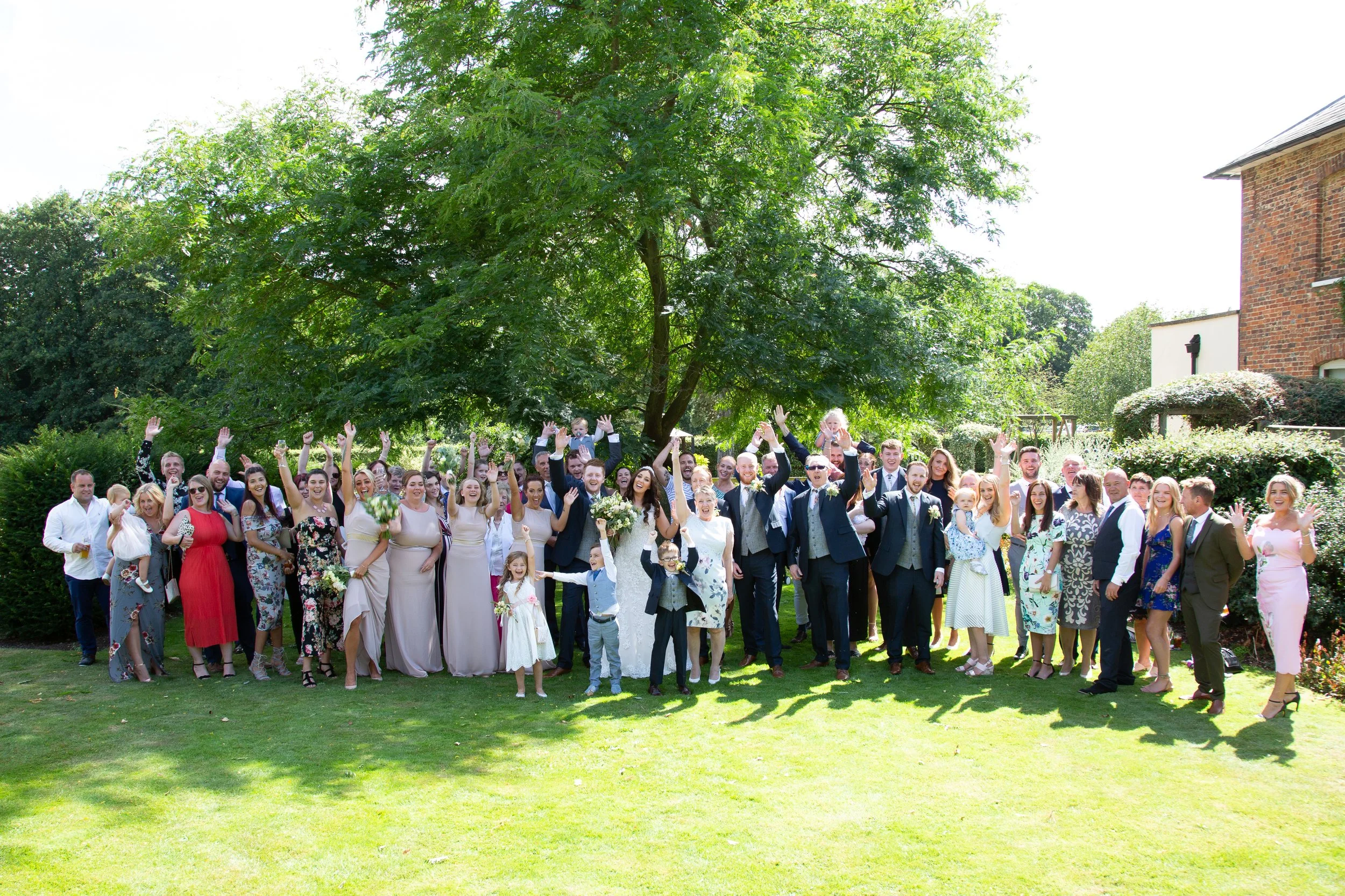 A large group of people dressed in formal and semi-formal attire celebrating outdoors on a sunny day, with a big green tree in the background.