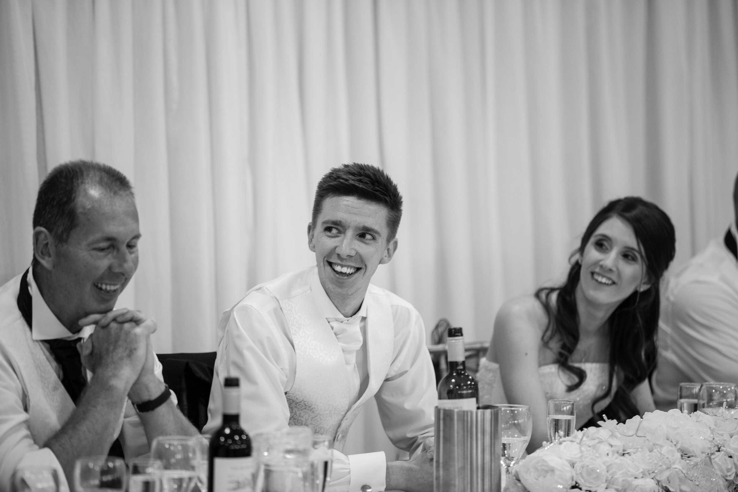A group of people at a wedding reception, including a smiling man in the center, a woman with dark hair on the right, and an older man on the left, sitting at a table with drinks and floral decorations.